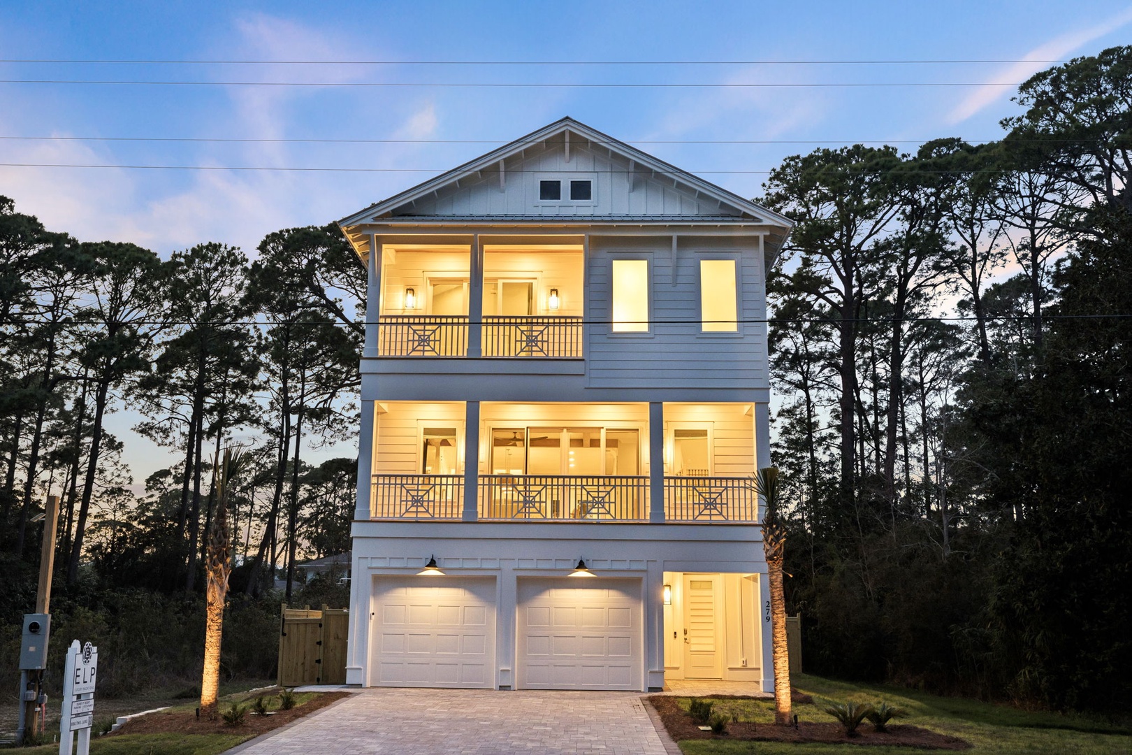 Beautiful three-story coastal home with warm lighting and covered balconies, nestled among mature trees in a peaceful residential setting.