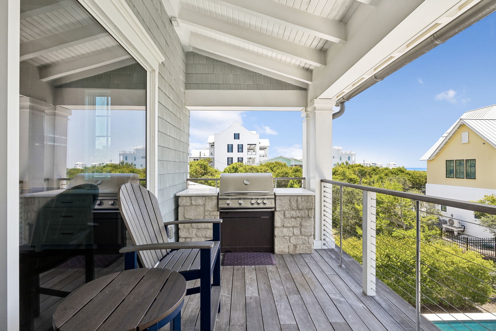 Spacious covered balcony features a built-in grill and relaxing seating, perfect for enjoying coastal breezes and views of lush greenery and nearby homes.