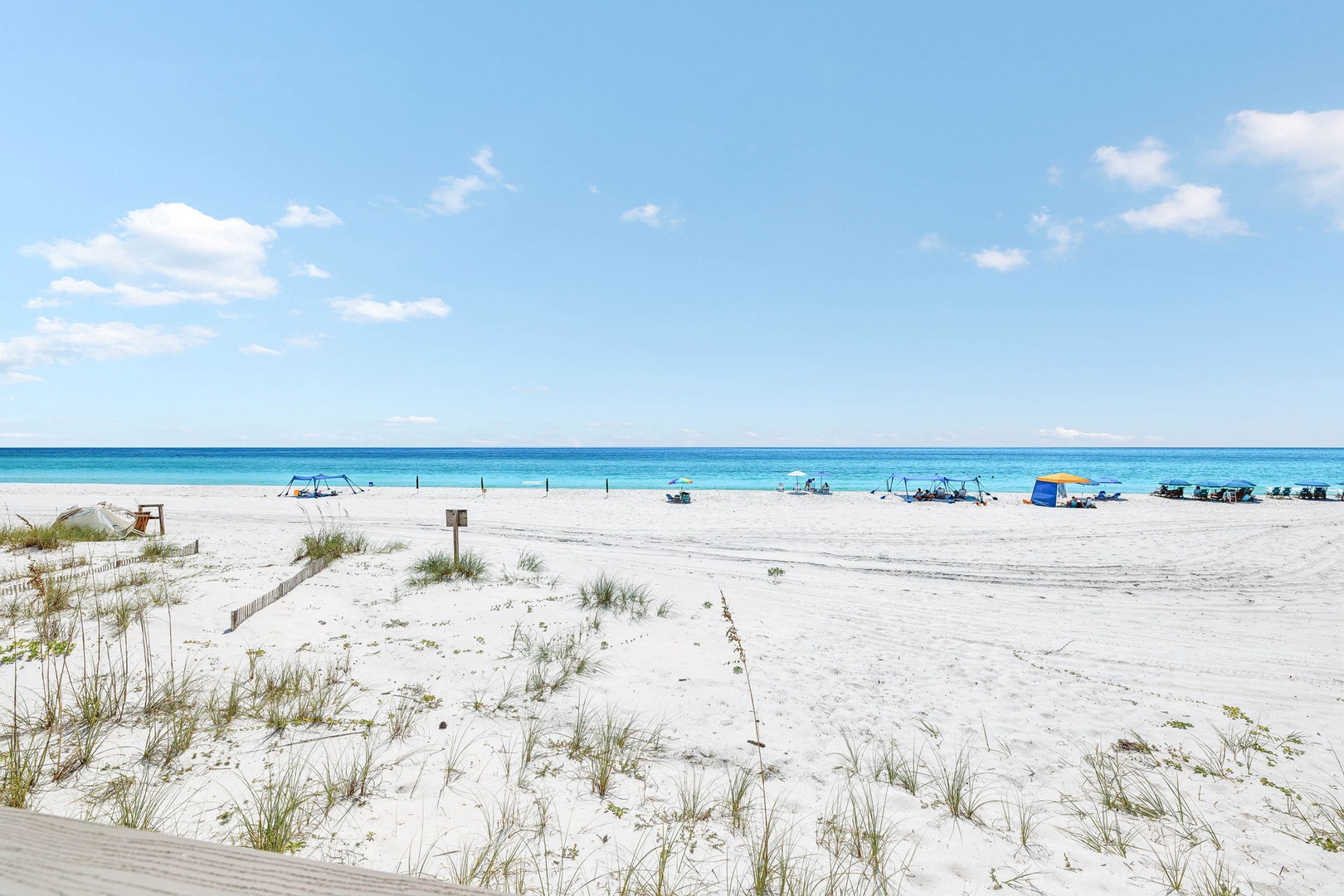 Stunning white sand beach with crystal-clear turquoise waters stretching to the horizon. Beach chairs and umbrellas dot the pristine shoreline under a bright sunny sky.