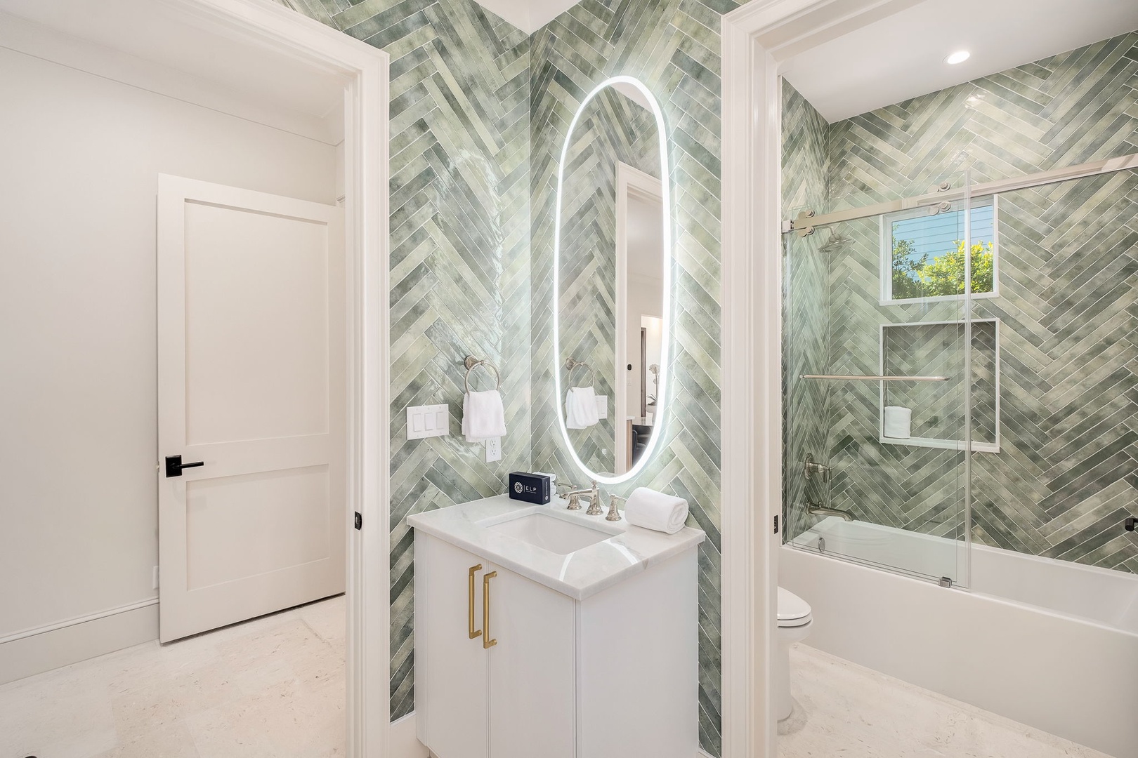 Admire the sophisticated herringbone tilework in calming green tones as you prepare for your day in this bright, contemporary bathroom featuring a sleek glass-enclosed tub and stylish LED mirror.