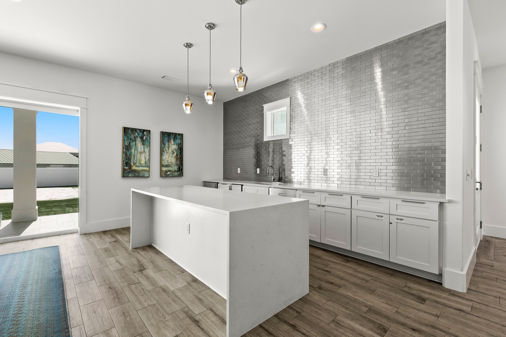A bright, modern kitchen features a sleek white island and metallic tile backsplash, with warm wood-look floors underfoot and lovely natural light streaming through the windows.