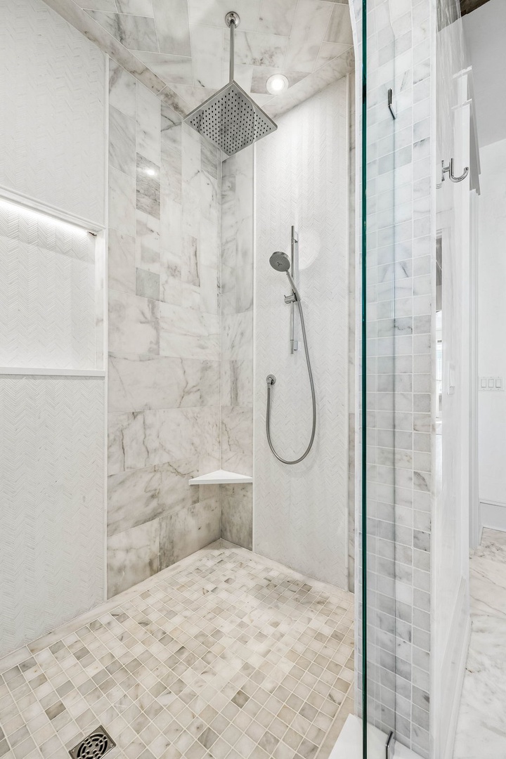 Step into a sleek, spa-like shower with luxurious marble walls, rainfall showerhead, and handheld spray. The built-in bench and bright lighting create a relaxing retreat for your mornings.