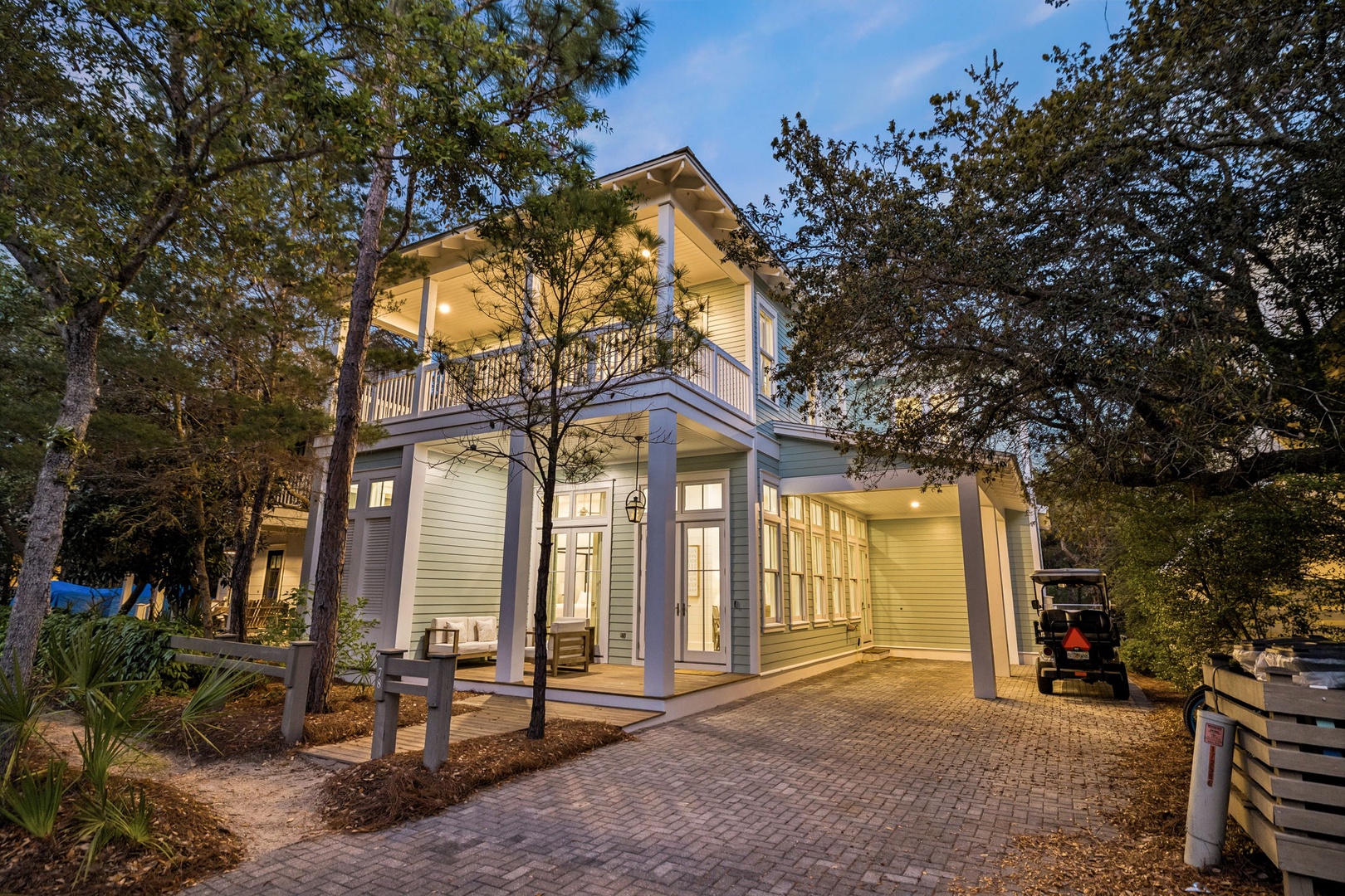 Charming coastal-style home nestled among towering trees with welcoming front porch and brick paver driveway for easy arrival.