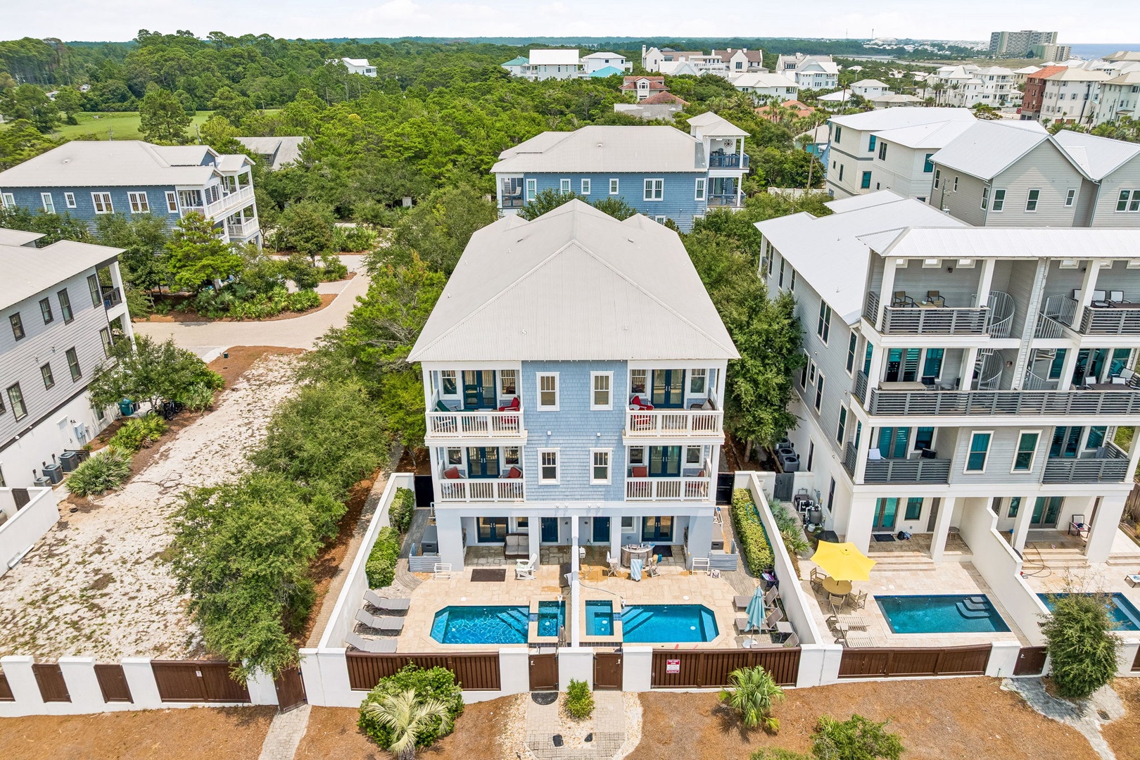 An aerial view of the property nestled in a coastal residential neighborhood, showcasing dual swimming pools and spacious balconies surrounded by lush greenery and nearby beach access.