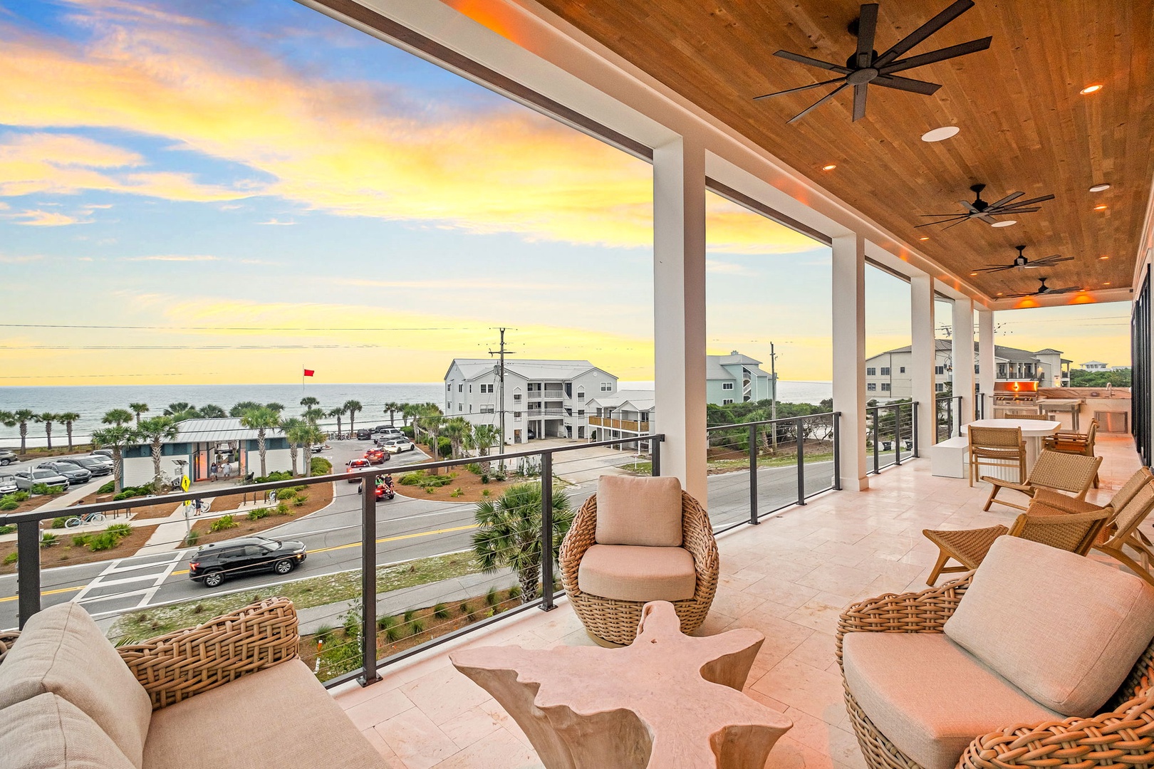 Modern covered balcony overlooking a coastal area at sunset, featuring comfortable wicker seating, ceiling fans, and views of palm trees and nearby beach buildings.
