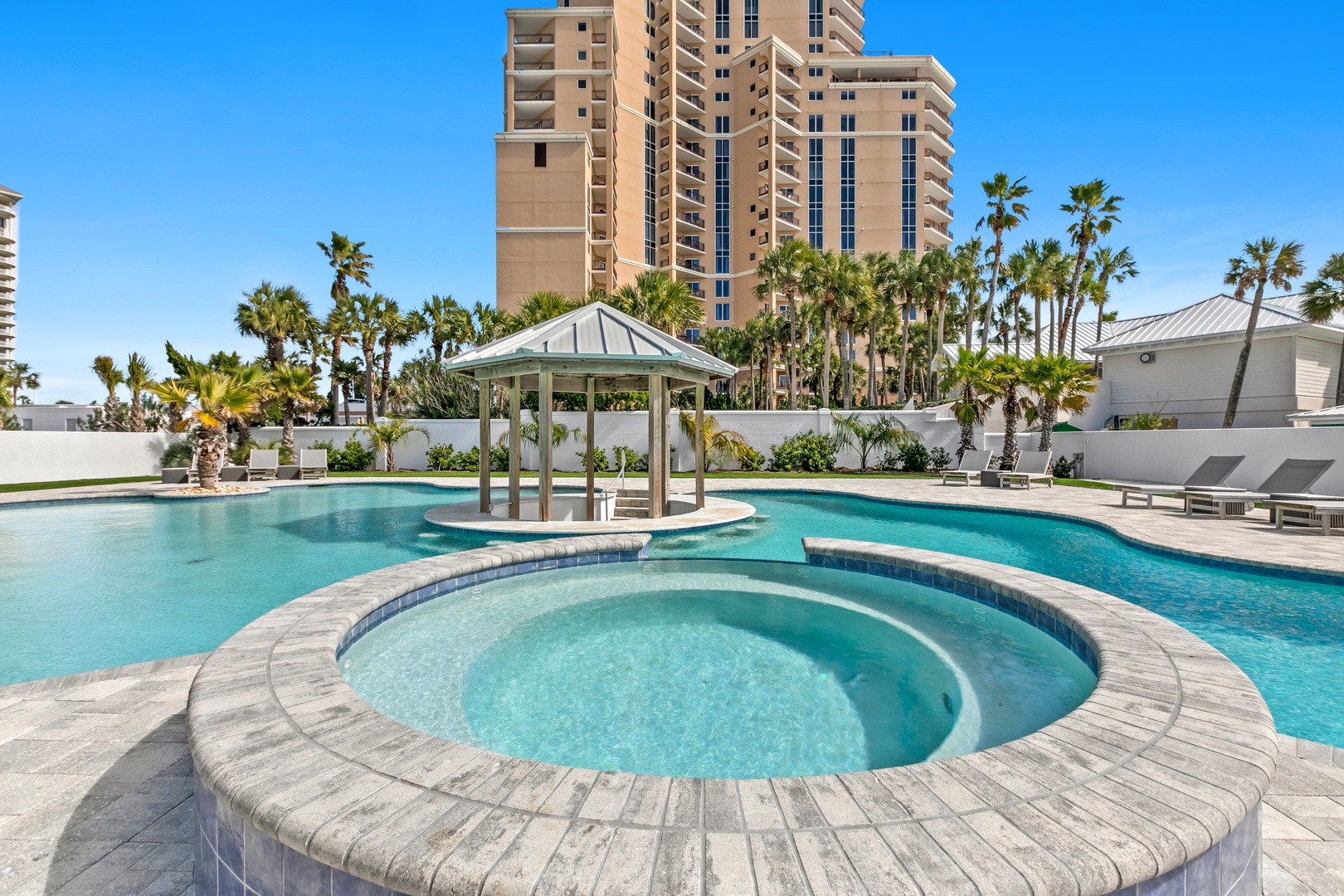 A large, resort-style pool with circular hot tub and shaded gazebo, surrounded by tropical palms and lounge chairs—your perfect spot for sunny relaxation.