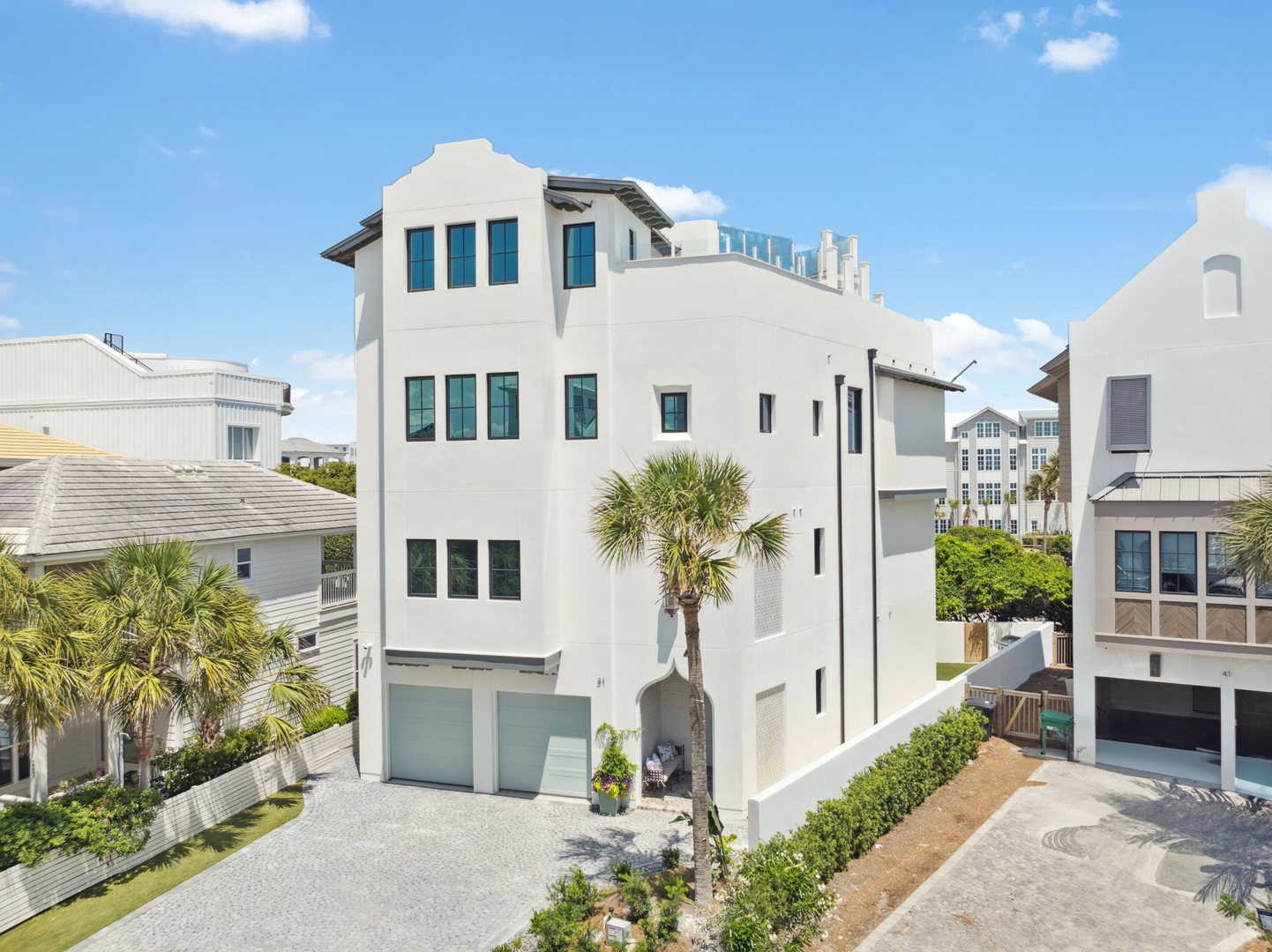 A striking white coastal property nestled among palm trees and beach homes, featuring classic architectural details and a rooftop terrace. The vacation rental stands prominently in this charming seaside neighborhood.