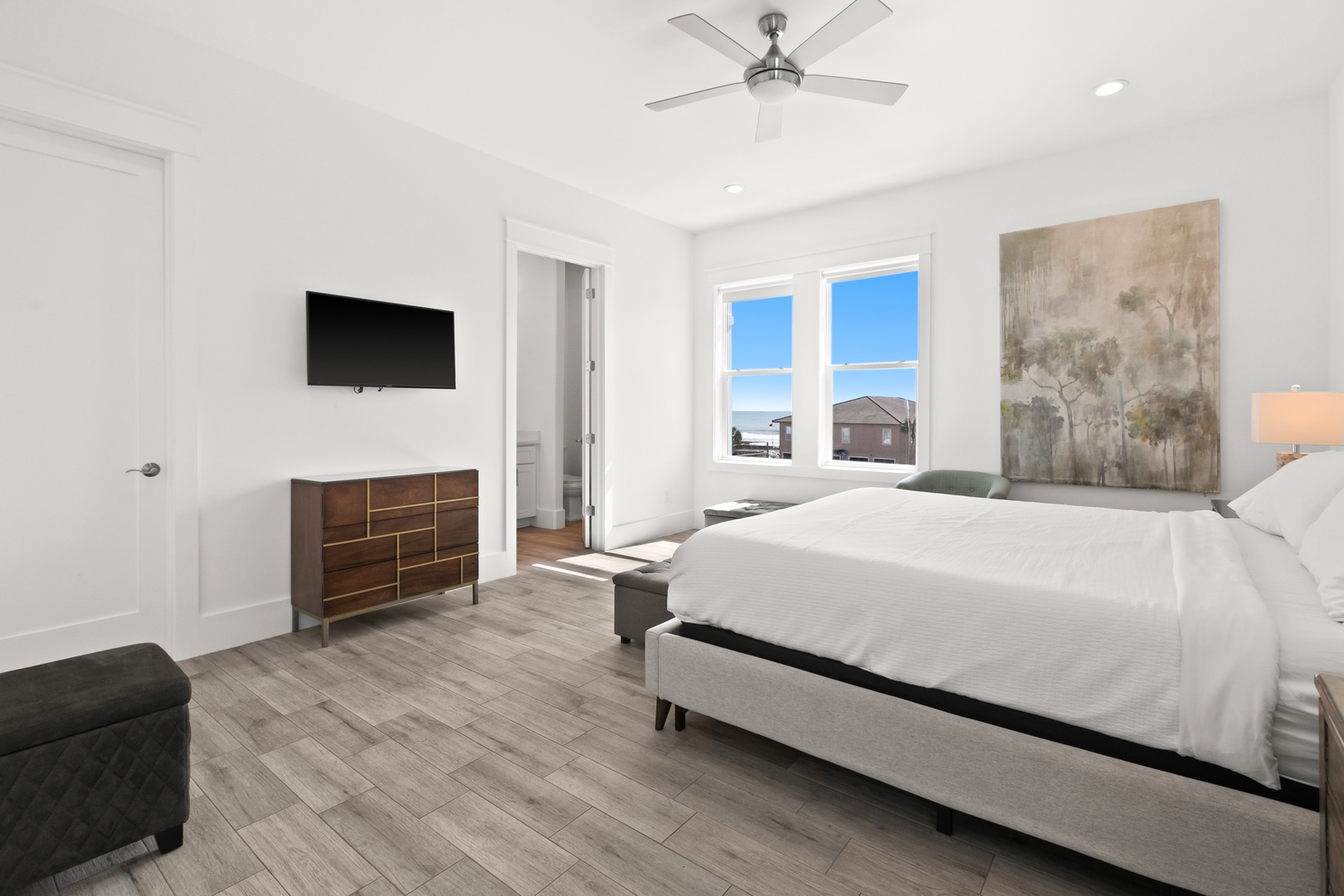 Your serene bedroom sanctuary features a plush bed, stylish furnishings, and a wall-mounted TV for your entertainment. Natural light floods through large windows, while a ceiling fan keeps you cool and comfortable.