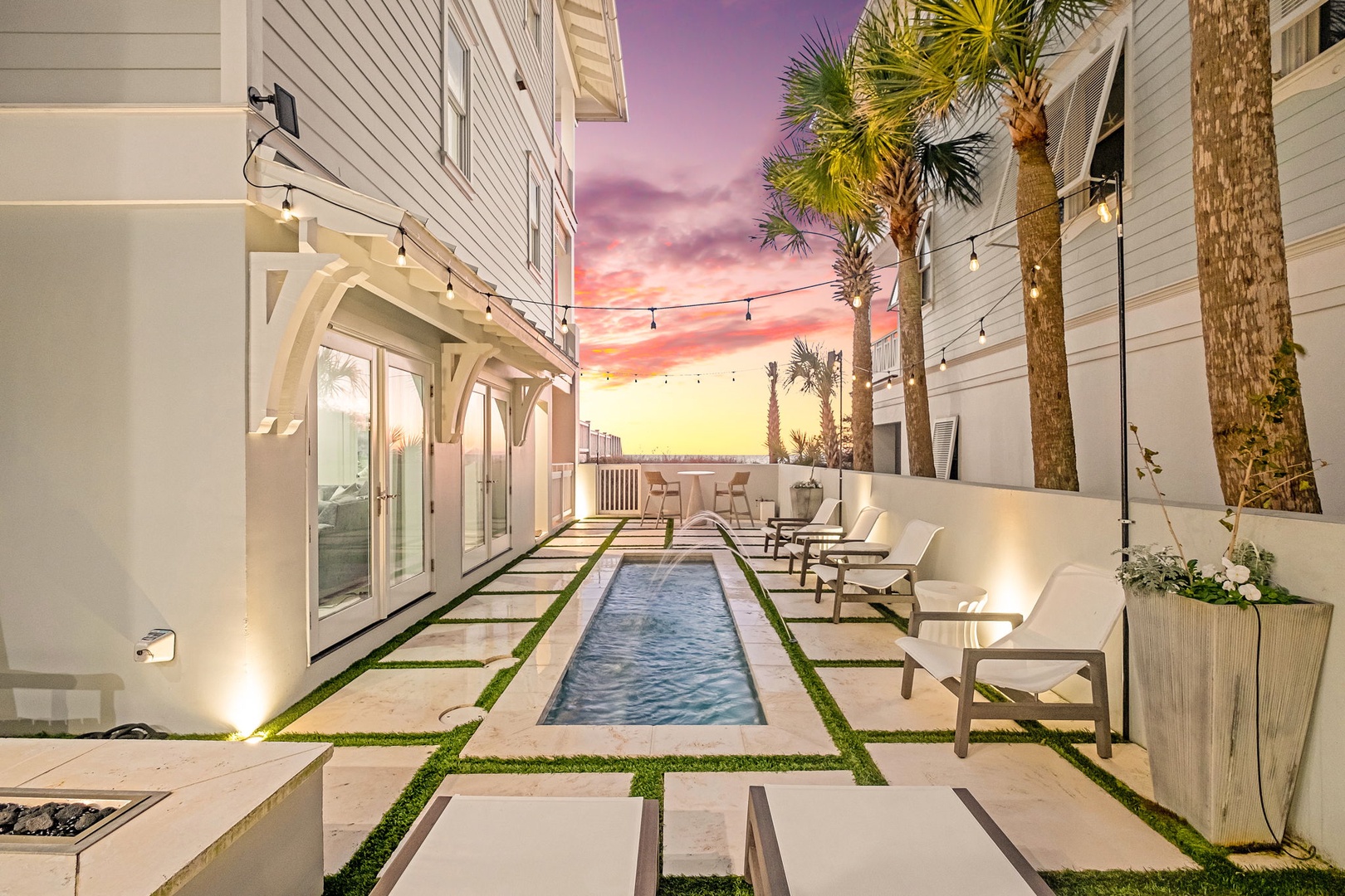 A stunning courtyard oasis captures the magical moment when day meets night, with palm trees silhouetted against a vivid purple-and-coral sky, while string lights and a sleek pool create an intimate retreat.