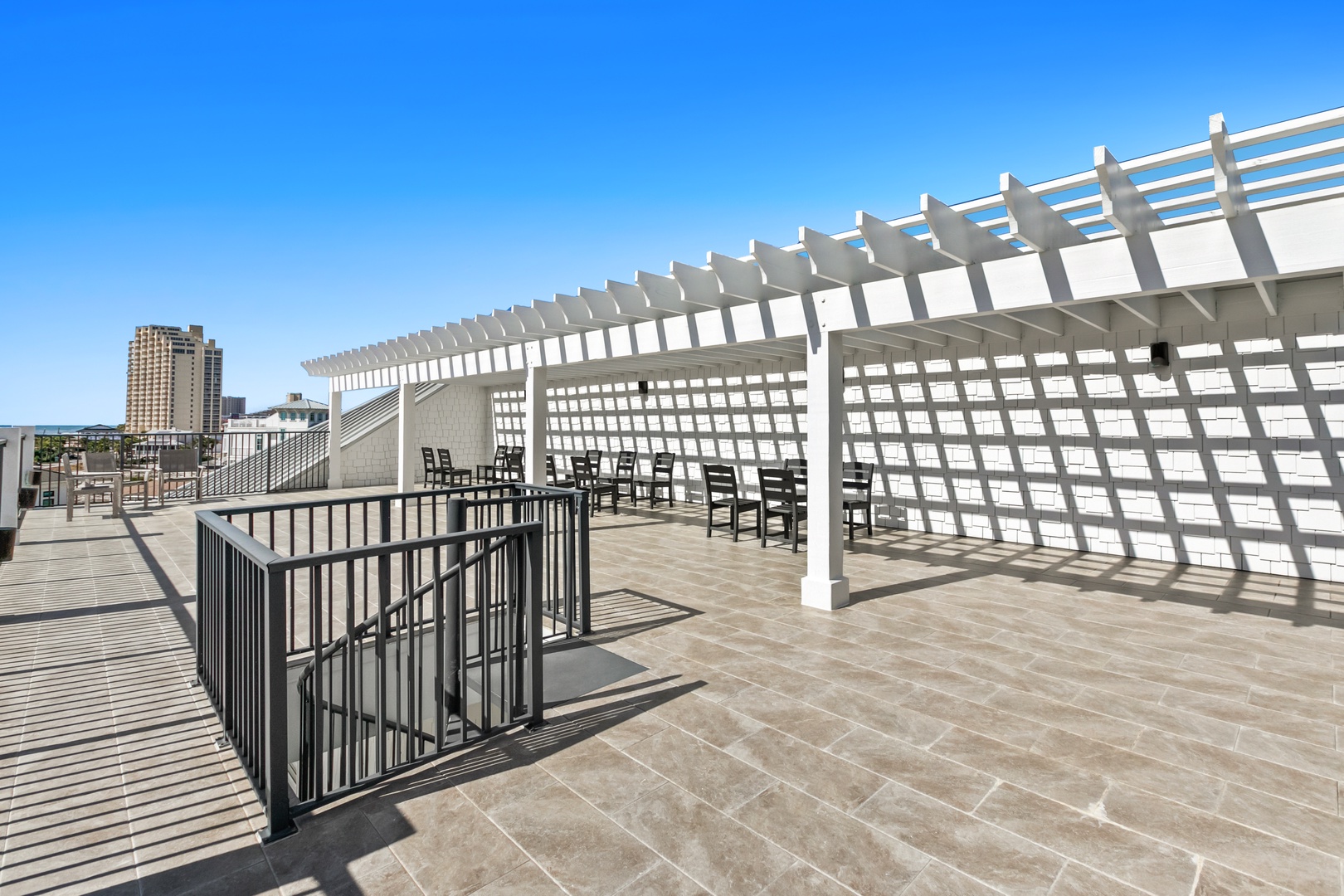 A bright rooftop terrace offers a modern pergola with seating and expansive views of the surrounding city skyline. The urban setting and clear blue skies create an inviting backdrop for this stylish outdoor communal space.