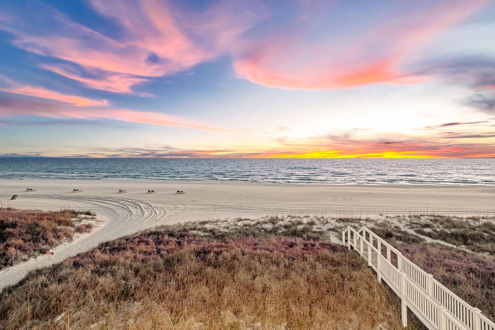 A stunning sunset paints the sky in brilliant shades of pink, orange, and gold over a pristine beach, with a sandy pathway and beach access leading down to the shore.