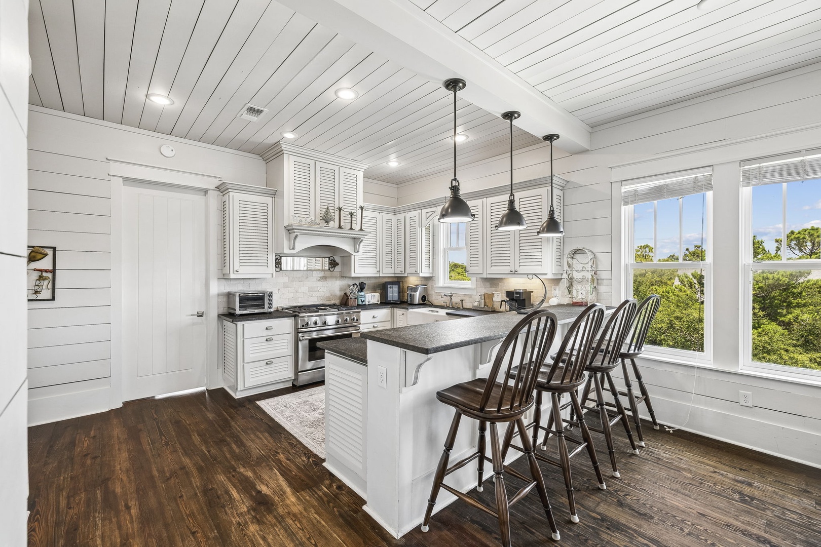 Your bright coastal kitchen features modern appliances and a generous island for gathering, framed by windows overlooking natural greenery.