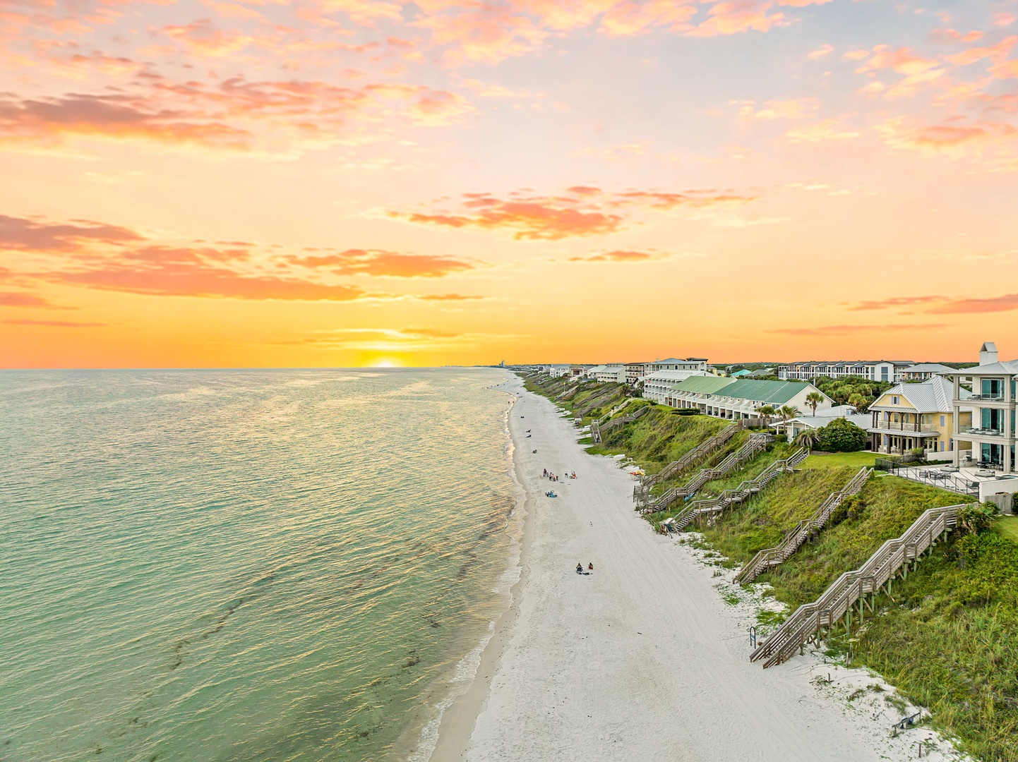 A stunning coastal sunset paints the sky in golden hues above a serene beachfront neighborhood where sandy shores meet calm waters.