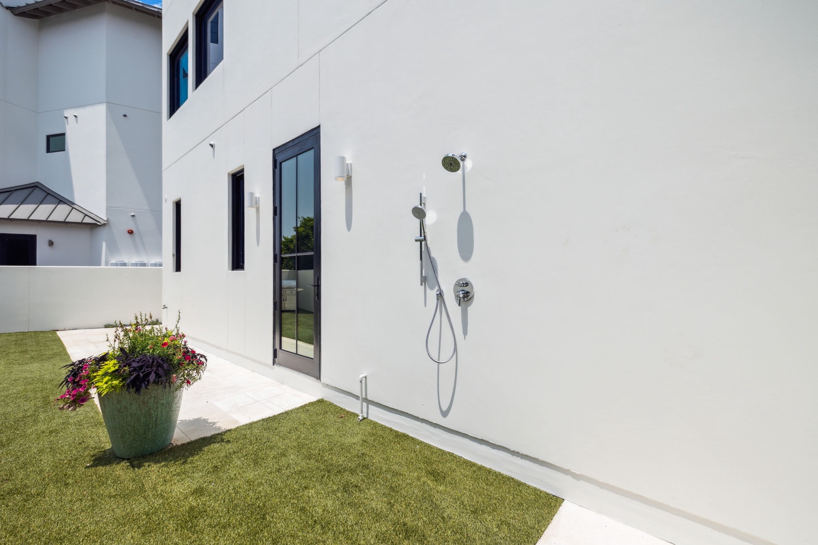 Modern two-story property features clean white exterior with black-framed glass door, outdoor shower fixture, and manicured lawn with colorful flower planter—a welcoming arrival area.