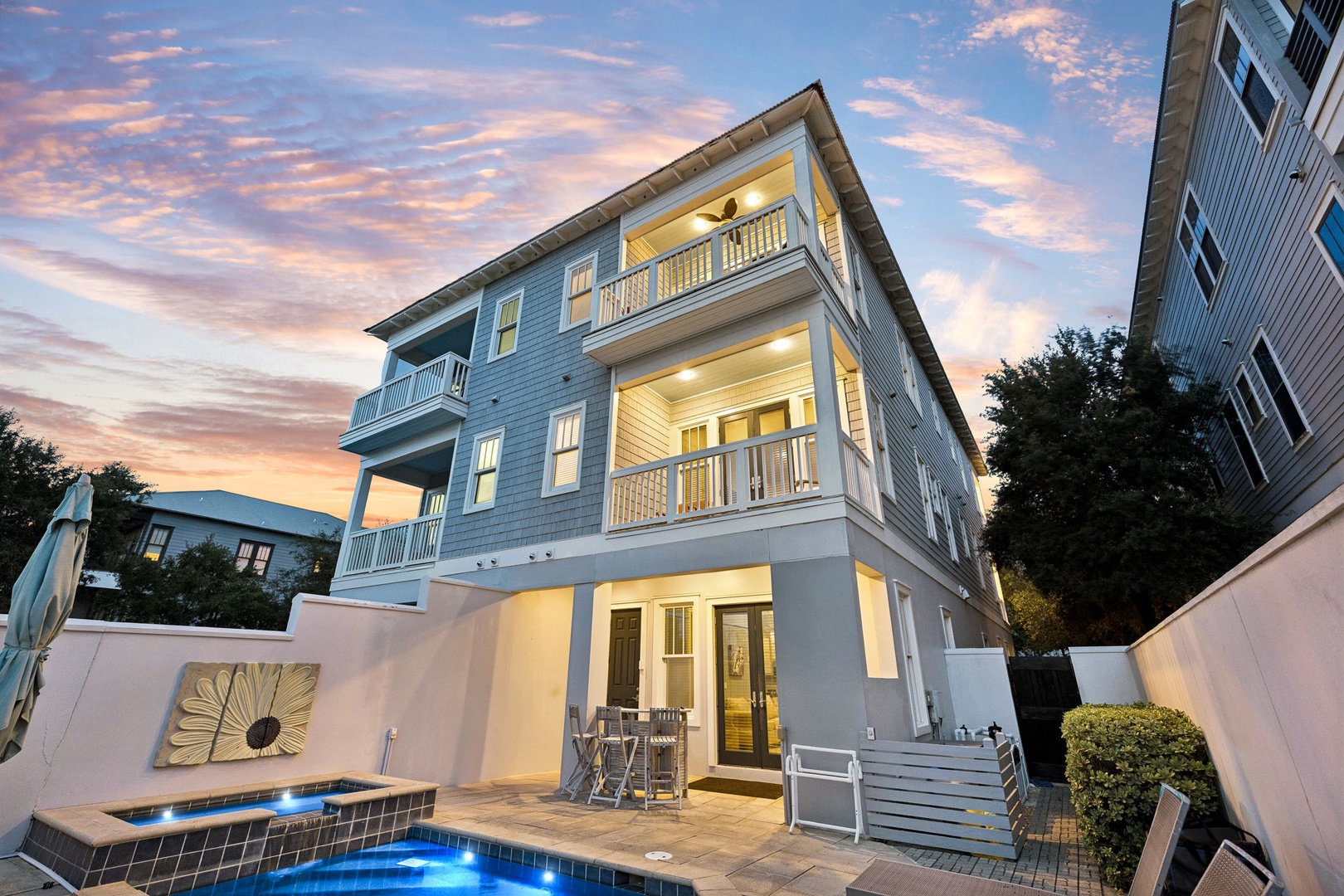 A three-story coastal home features private balconies, a sparkling pool with warm evening lighting, and an inviting outdoor patio—perfect for relaxing after a day at the beach.