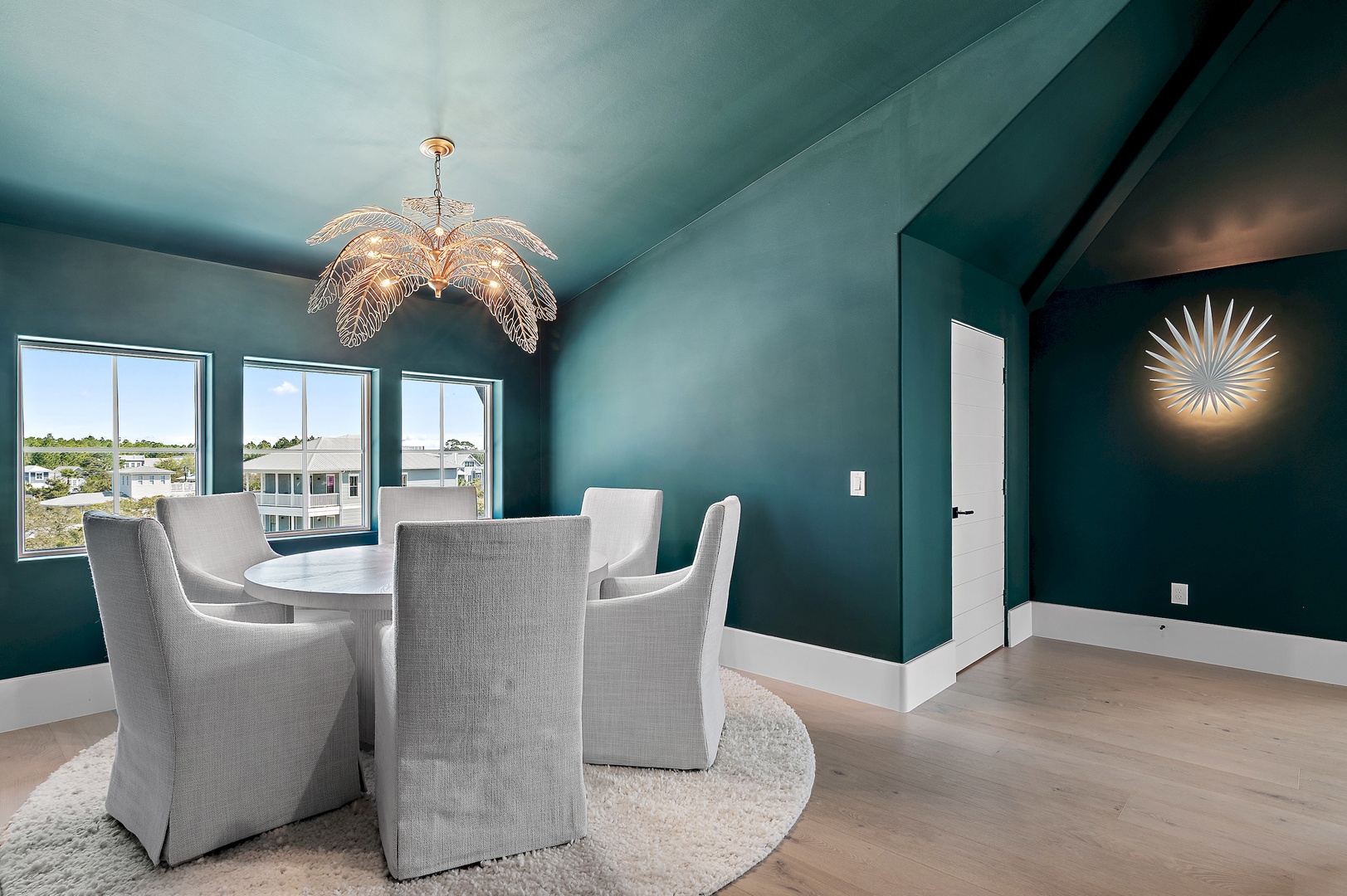 Gather around the elegant dining table beneath a stunning statement chandelier, where rich teal walls and natural light create an inviting space for memorable meals together.