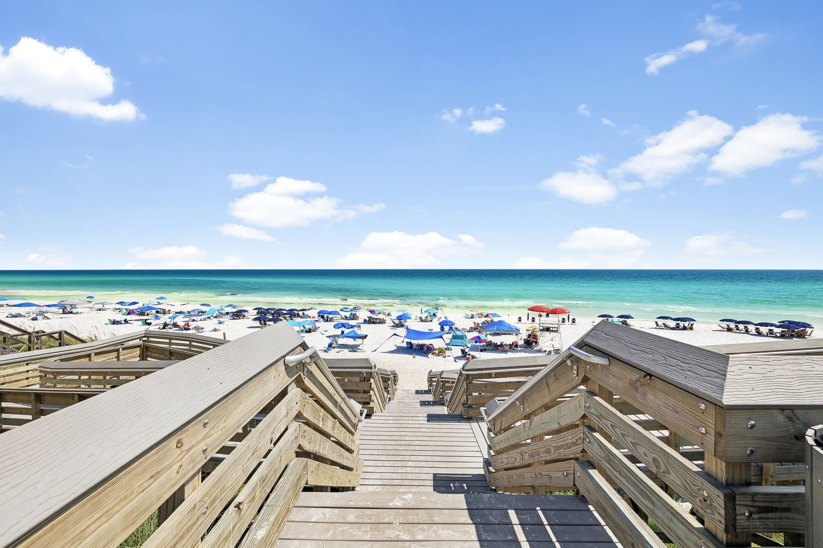 Wooden stairs lead down to pristine white sand and turquoise waters where your beach day awaits. Colorful umbrellas dot the shore, promising shade and relaxation just steps from the surf.