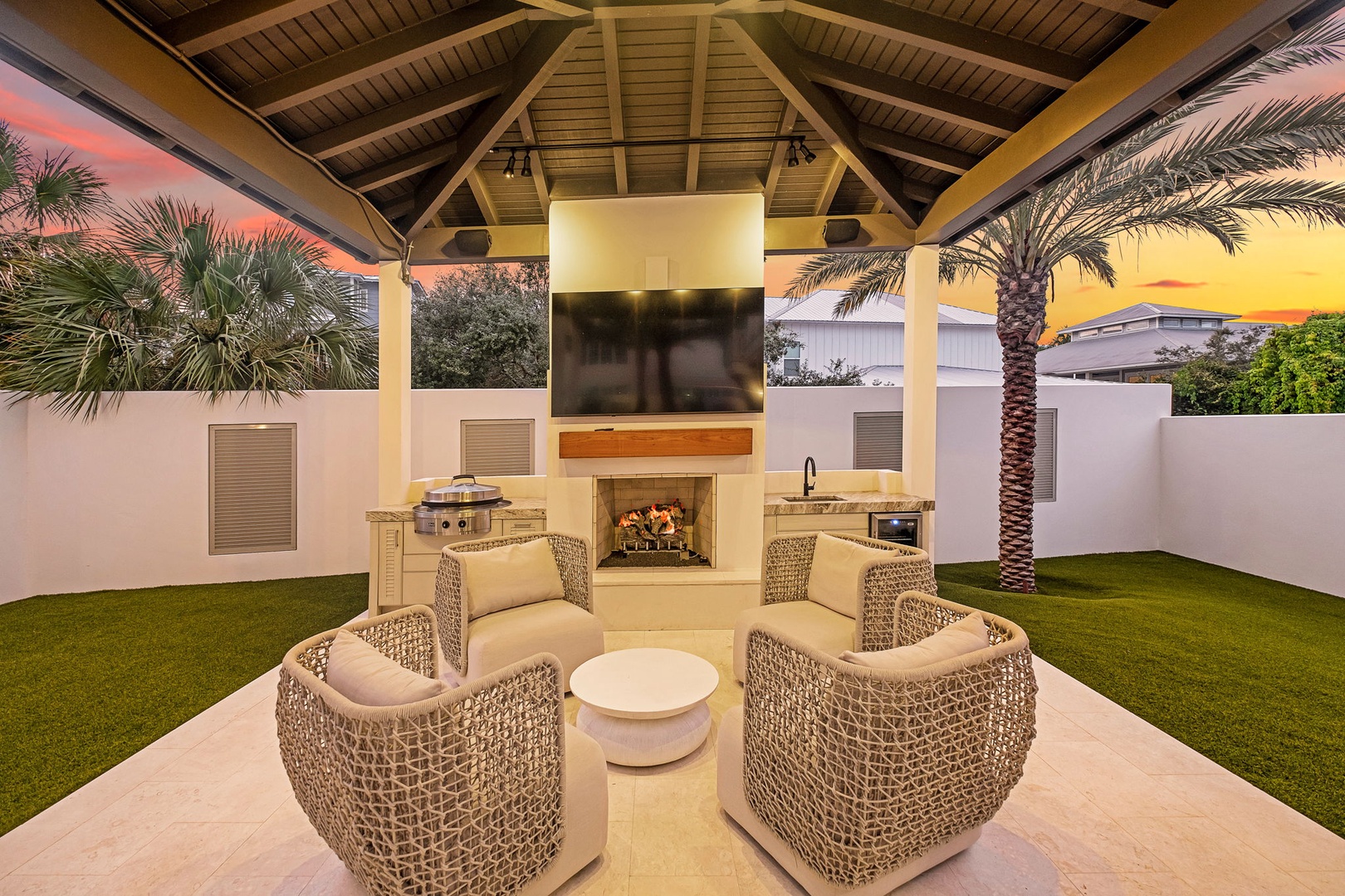 Unwind in your covered outdoor lounge with a cozy fireplace, comfortable seating, and a stunning sunset backdrop framing the palms—perfect for evening gatherings or quiet relaxation.