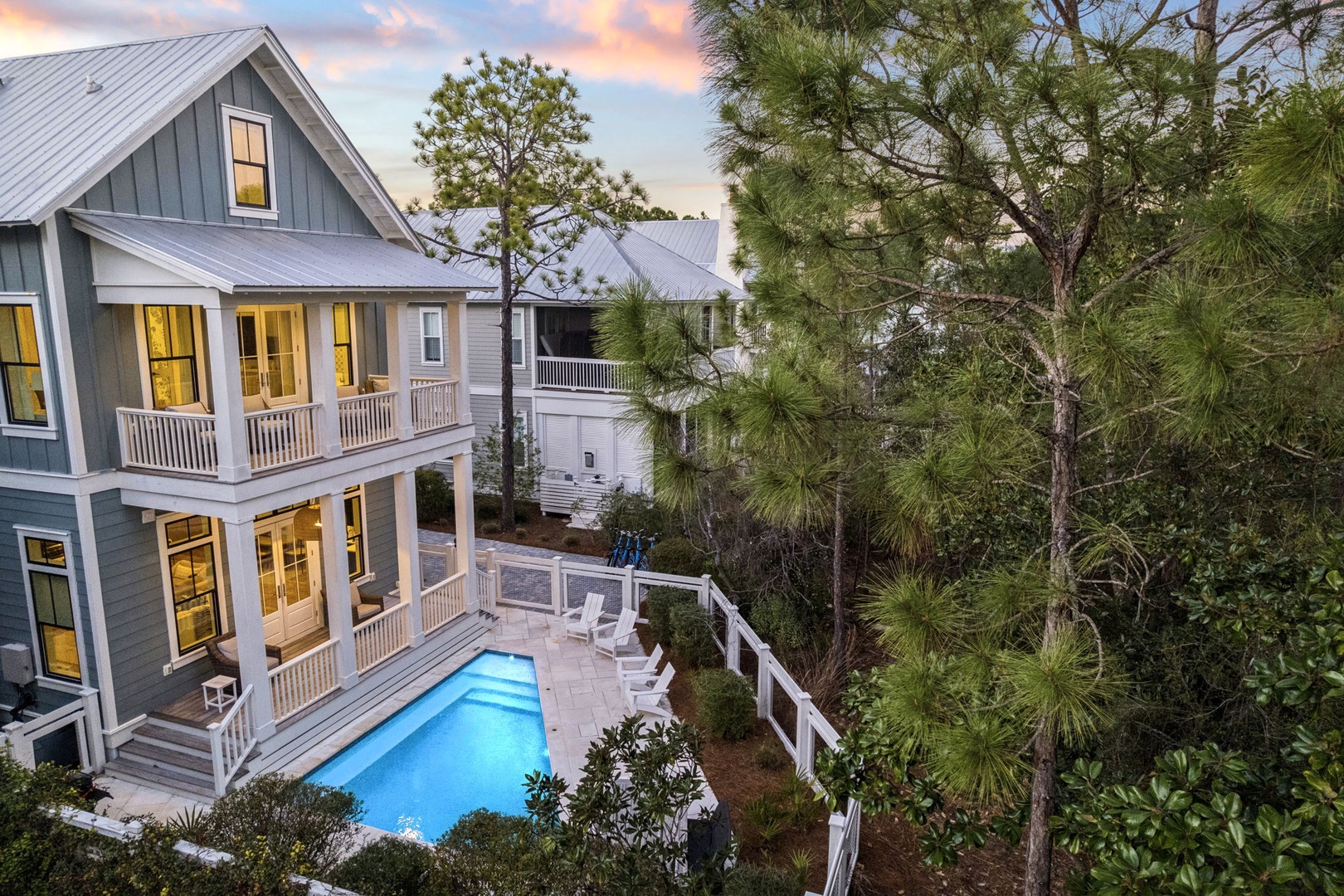 Charming coastal home with private pool nestled among towering pines, featuring wraparound porches and warm evening lighting.