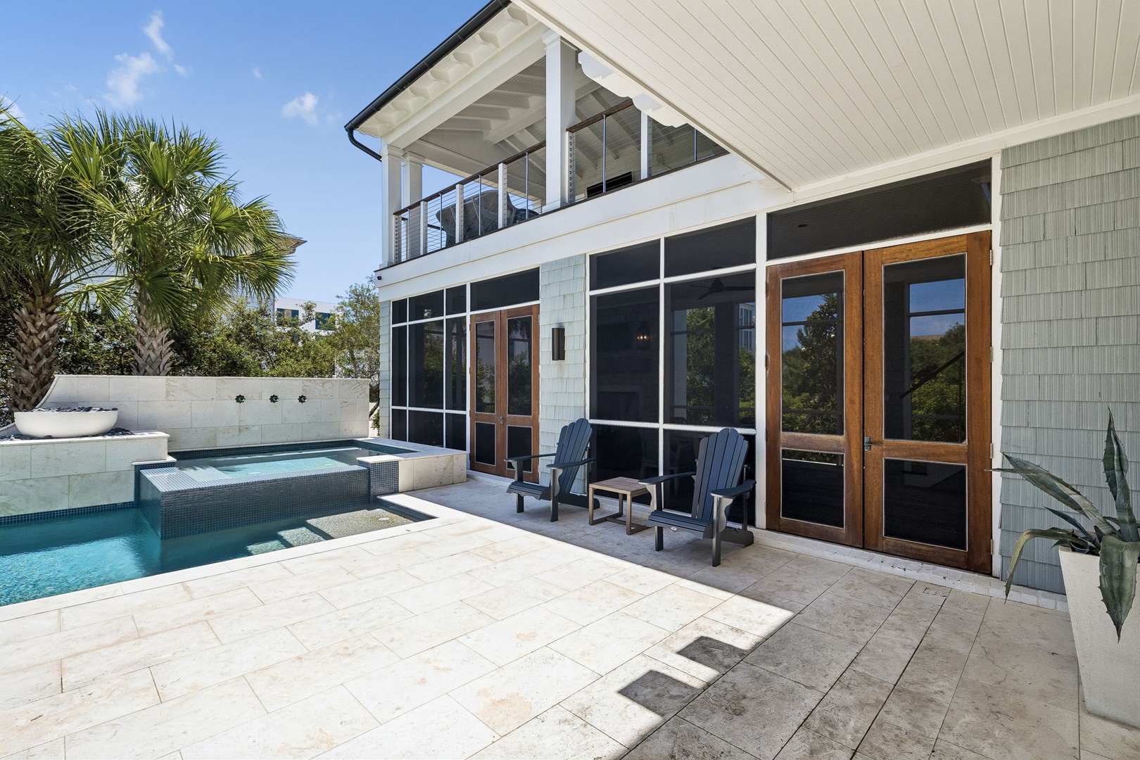 Modern coastal home featuring a sparkling pool, screened porch with seating, and second-story balcony, all set among tropical palms beneath a bright blue sky.