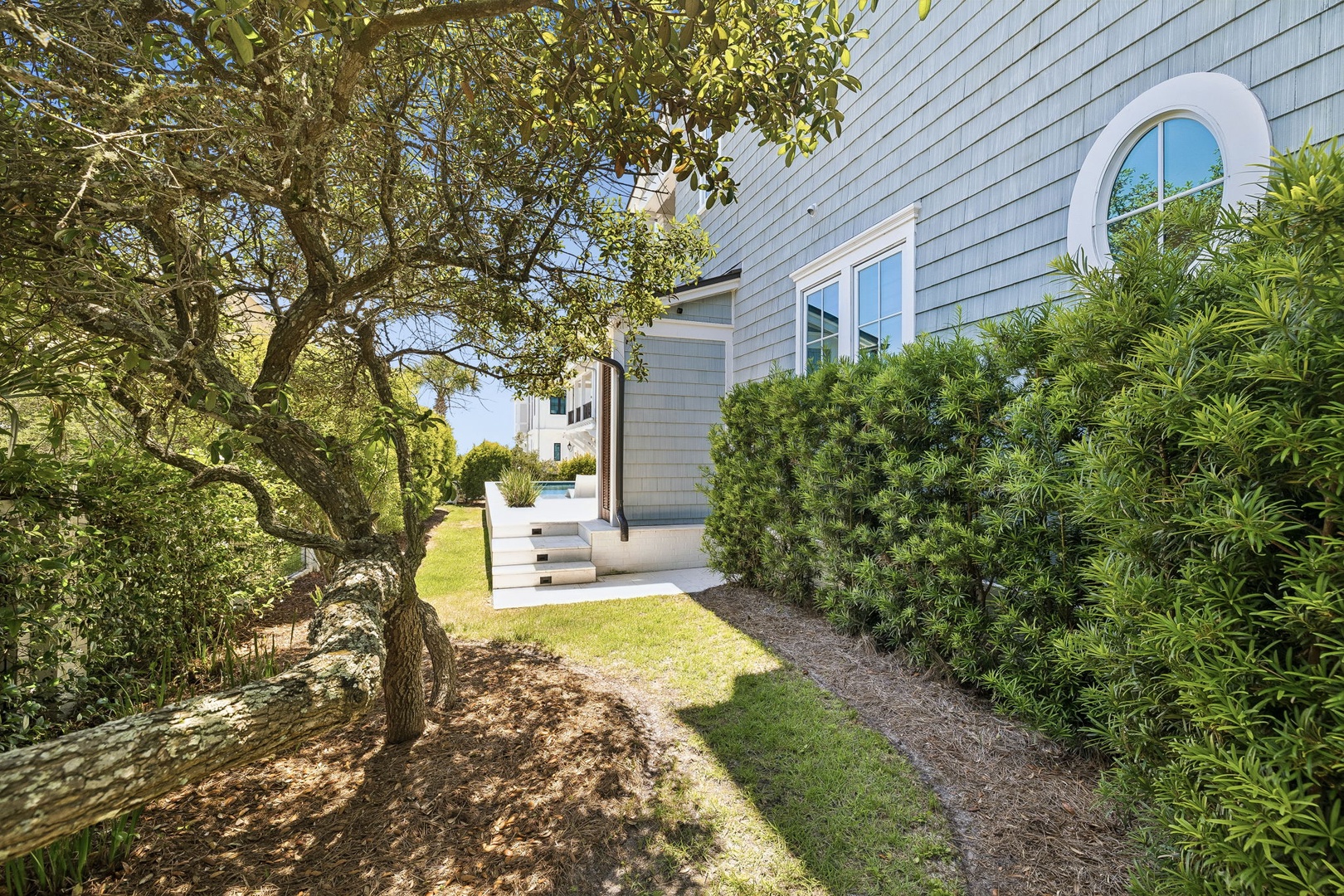 A charming coastal-style entrance nestled among mature trees and lush greenery, with a distinctive arched window accent marking the arrival to this inviting home.