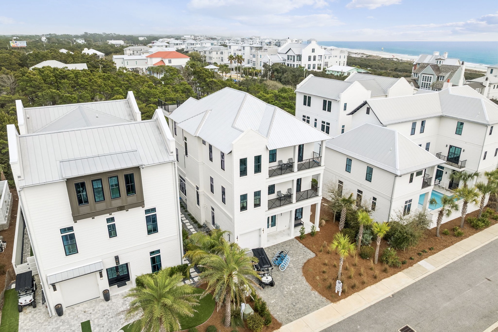 Aerial view of coastal vacation homes with white architecture and metal roofs, nestled among palm trees near the beach.