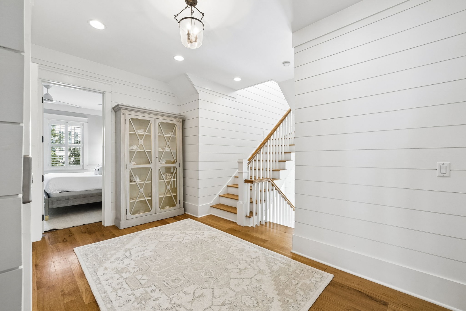 Natural light streams through the open door, inviting you into this charming hallway with crisp shiplap walls, warm hardwood floors, and elegant touches like the glass-front cabinet. The staircase beckons you to explore your upstairs retreat.