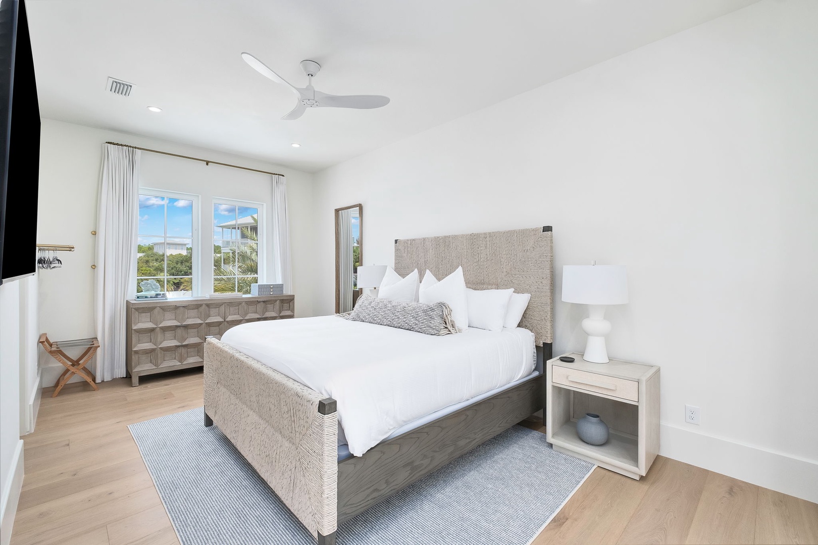 Slip into crisp white linens in your serene coastal bedroom. Natural light streams through windows framing palm trees and sunshine. Gentle ceiling fan and thoughtful furnishings invite you to unwind completely.