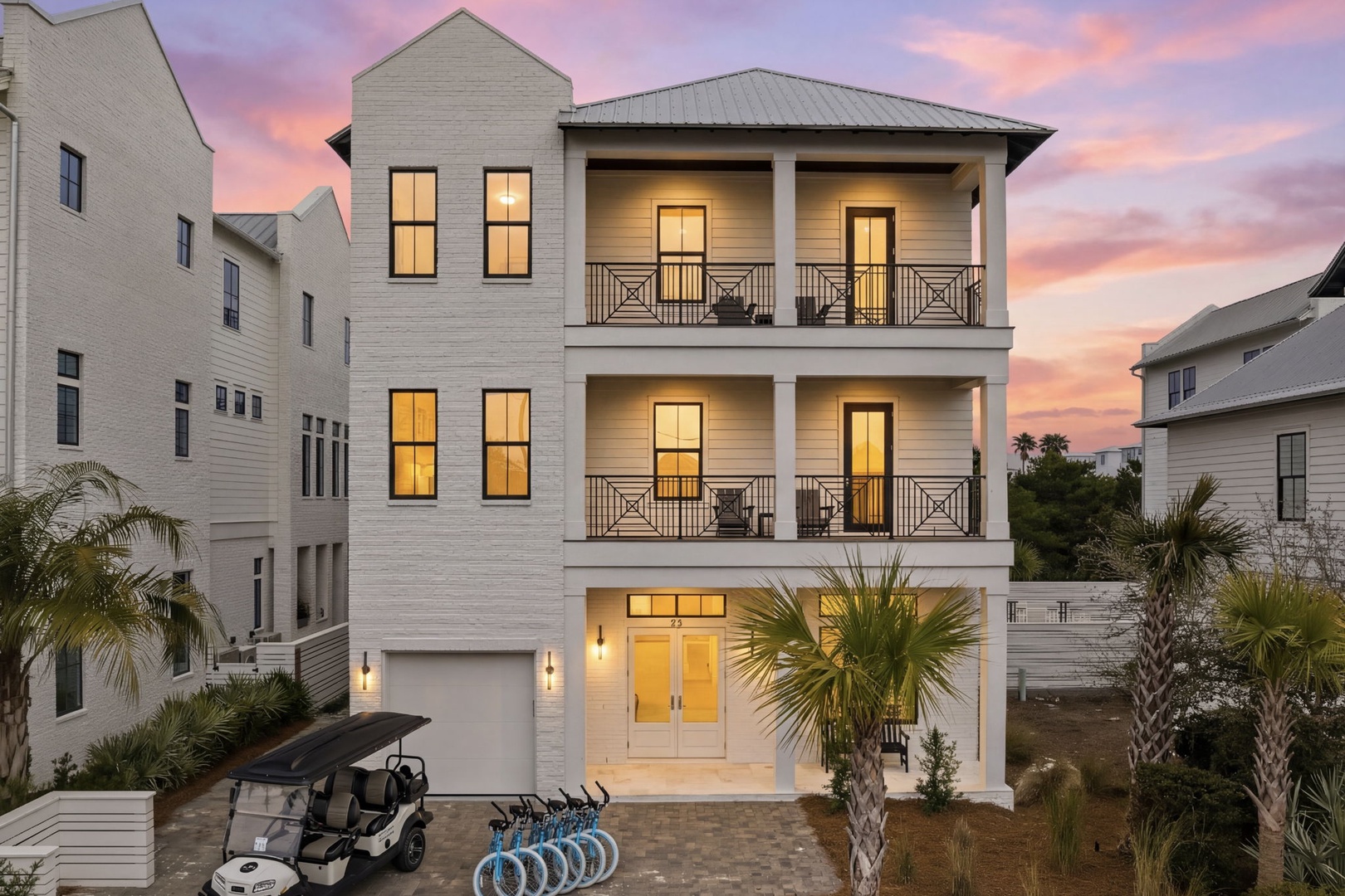 A stunning three-story coastal home with double balconies and warm lighting, nestled among palm trees in a charming beach community.