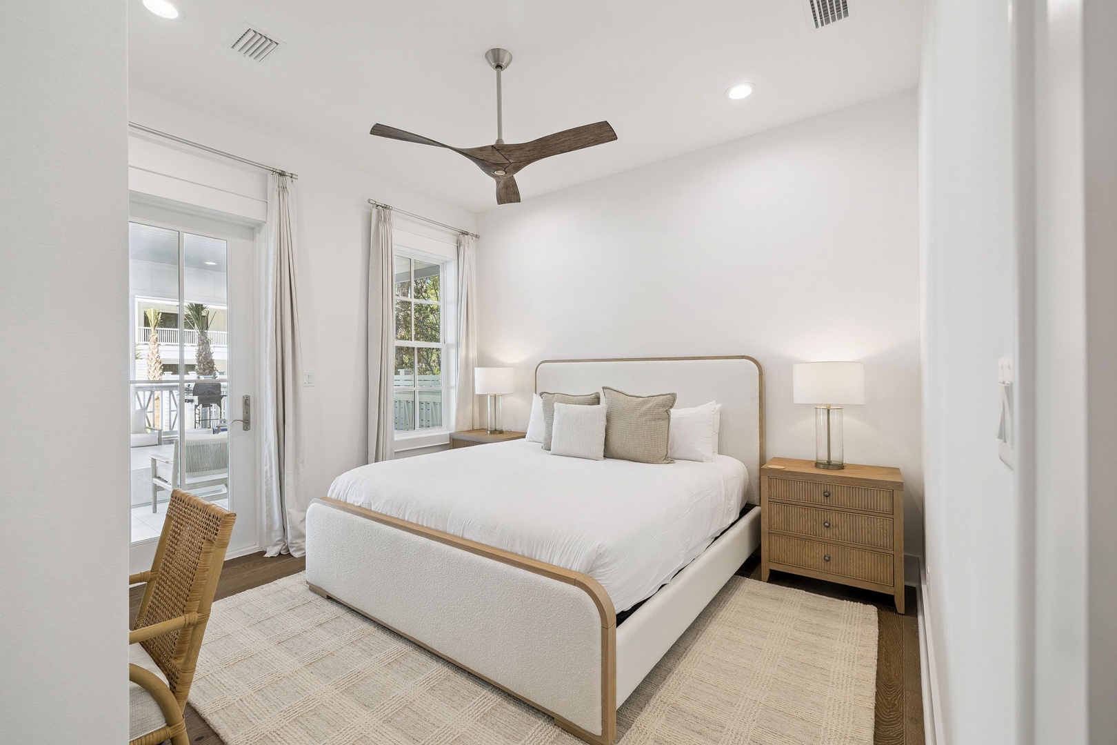 Sink into crisp white linens in your serene bedroom, where natural light streams through windows and coastal breezes from the ceiling fan create perfect sleeping comfort.