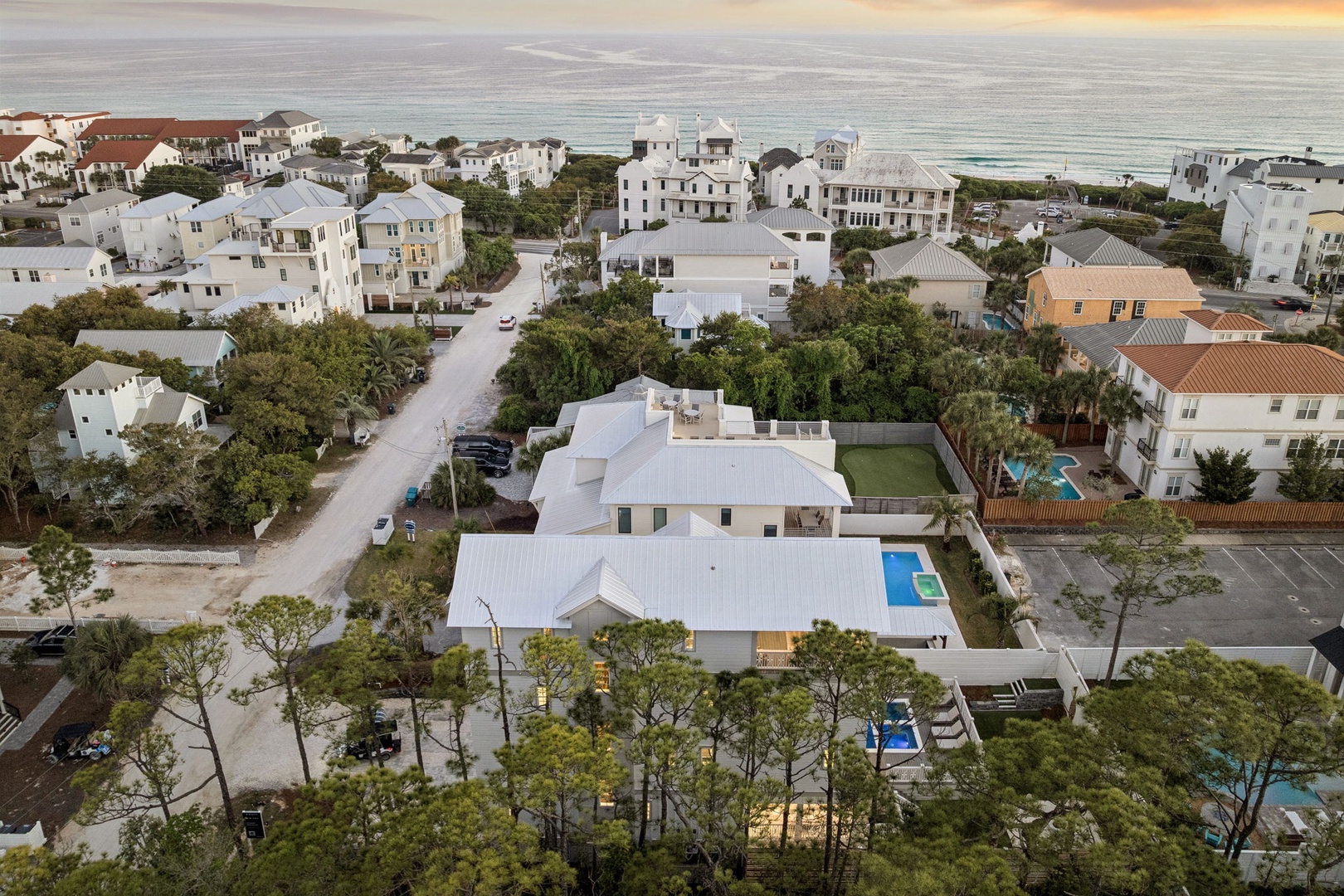 Aerial view of a coastal residential area with vacation homes, swimming pools, and tree-lined streets near the beach.
