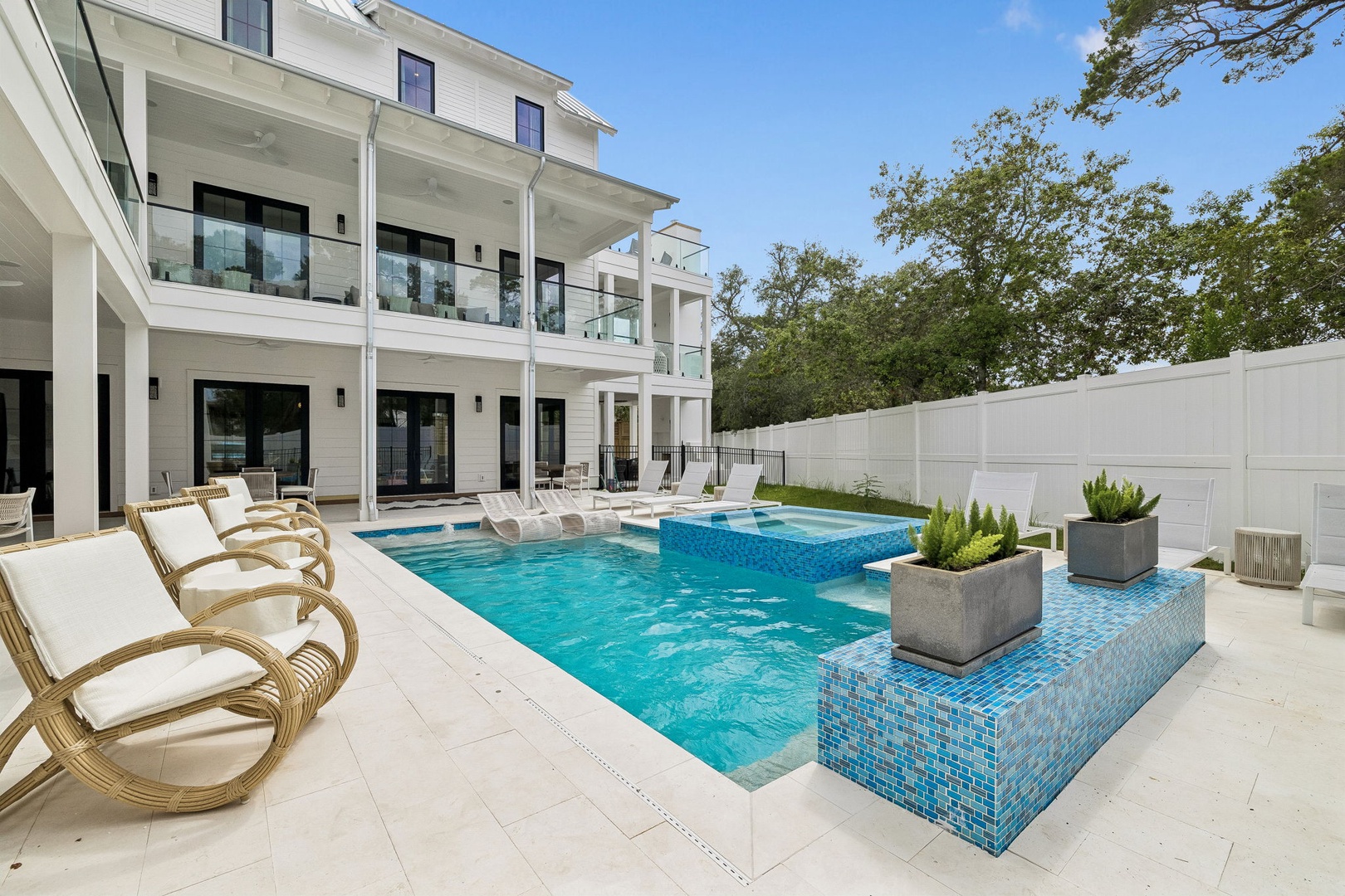 Modern coastal retreat with stunning pool featuring mosaic-tile water features and lounge areas surrounded by lush trees and white privacy fencing. Perfect for peaceful outdoor living.