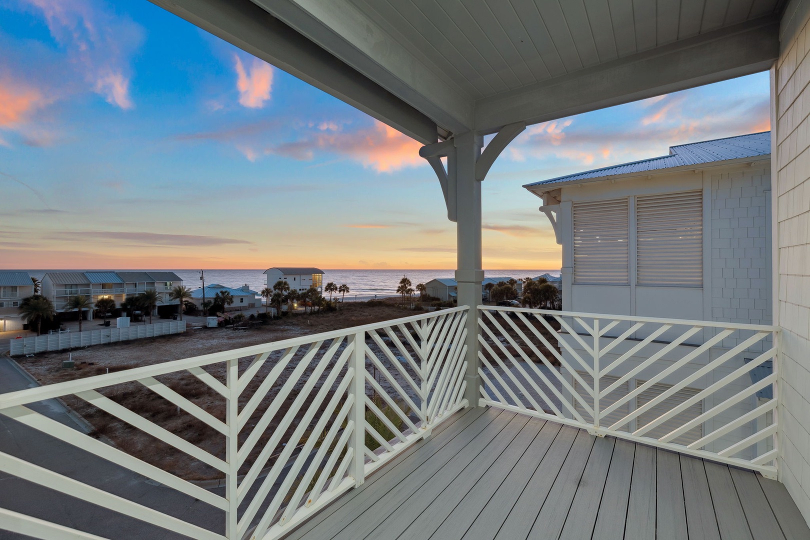 Covered balcony overlooking coastal homes and palm trees with ocean views during golden hour sunset.