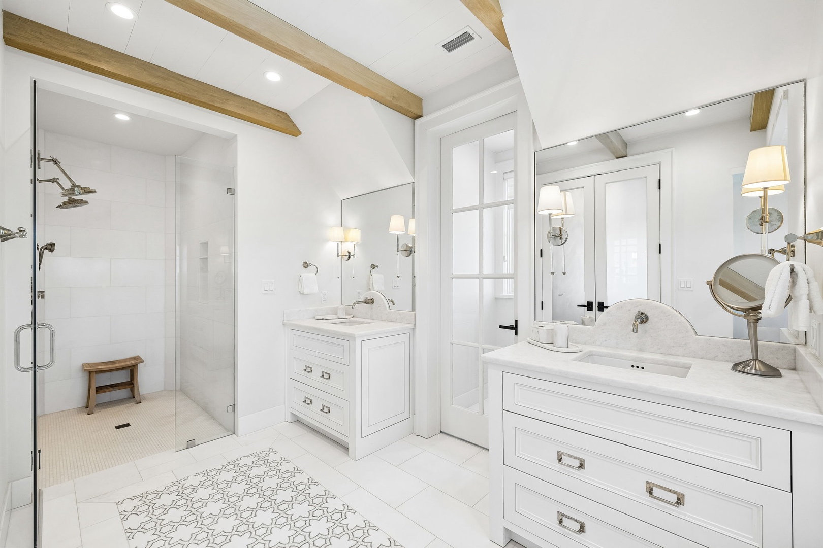 Step into your serene private bathroom, where crisp white finishes meet warm wood beams overhead. The spacious walk-in shower and elegant dual vanities with marble countertops create a spa-like retreat for your morning routine.