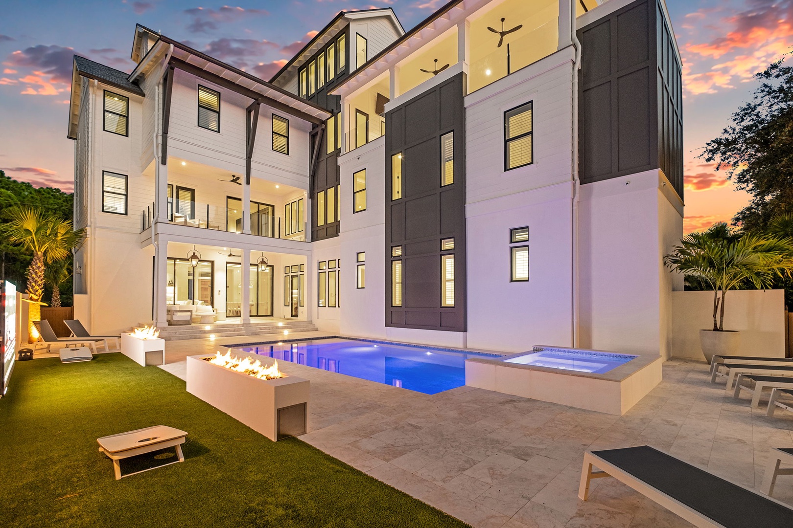Stunning modern architecture with sleek white and charcoal façade. The backyard oasis features a pool, fire feature, spa, and tropical palms under a vibrant sunset sky.