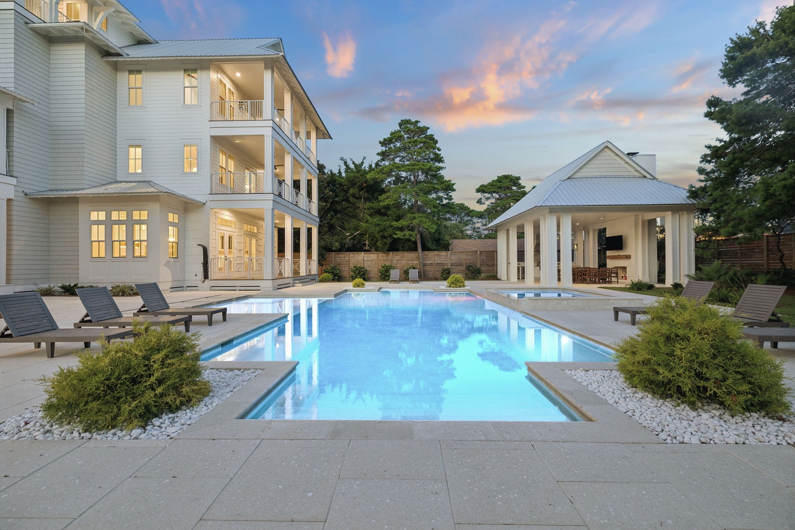A stunning white coastal-style property featuring an inviting pool and cabana pavilion, set under a dramatic evening sky with landscaped grounds and modern outdoor living spaces.