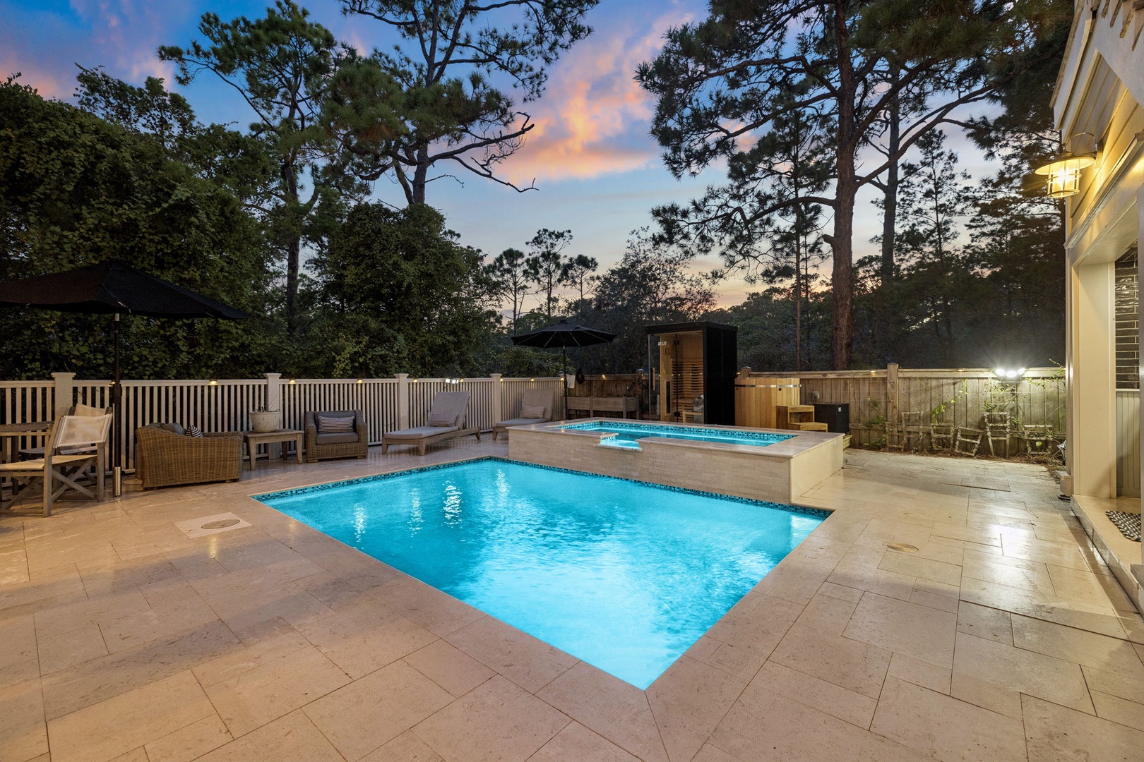 The property features a private outdoor pool and hot tub surrounded by tall pines, with warm lighting creating an inviting ambiance beneath a beautiful dusk sky.
