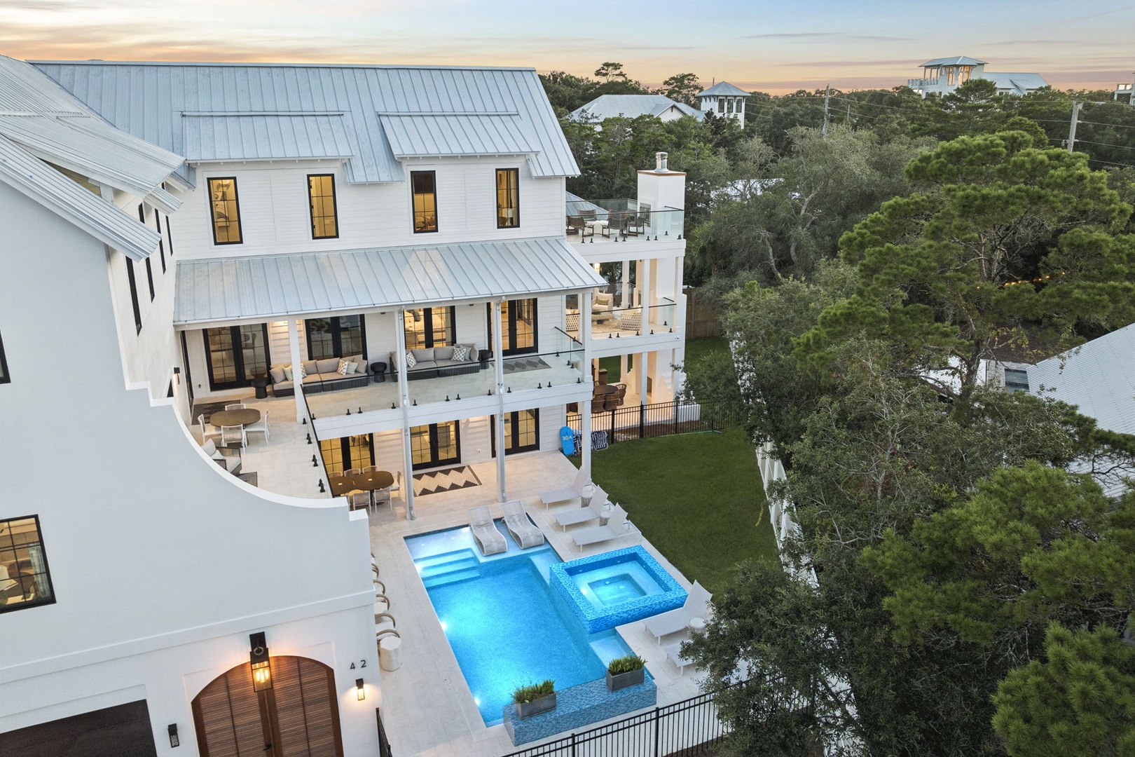 A striking coastal property featuring pristine white architecture, multiple private balconies, and a sparkling pool surrounded by lush natural greenery as the sun sets over the treetops.