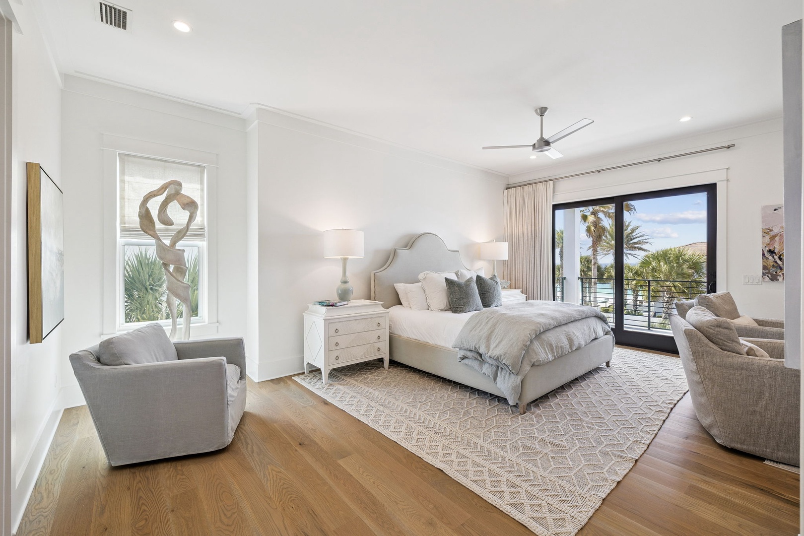 Relax in your serene bedroom featuring elegant coastal decor, plush seating, and French doors opening to peaceful palm views.