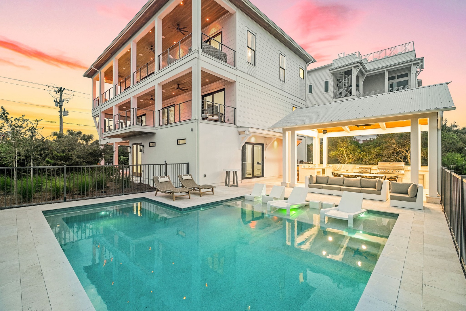 A stunning contemporary property featuring a private pool and covered outdoor living space, captured during a vibrant pink and orange sunset. The modern three-story building showcases clean architectural lines and multiple balconies.