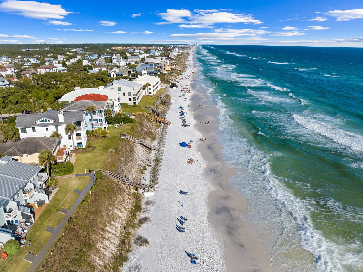 This aerial view captures a stunning coastal community nestled between turquoise waters and pristine white sand beaches, surrounded by natural beauty and charming beachfront properties.