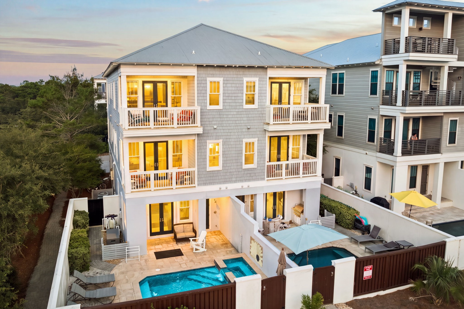 A stunning three-story coastal retreat featuring a private pool, multiple balconies, and lush greenery overlooking the neighborhood at sunset.