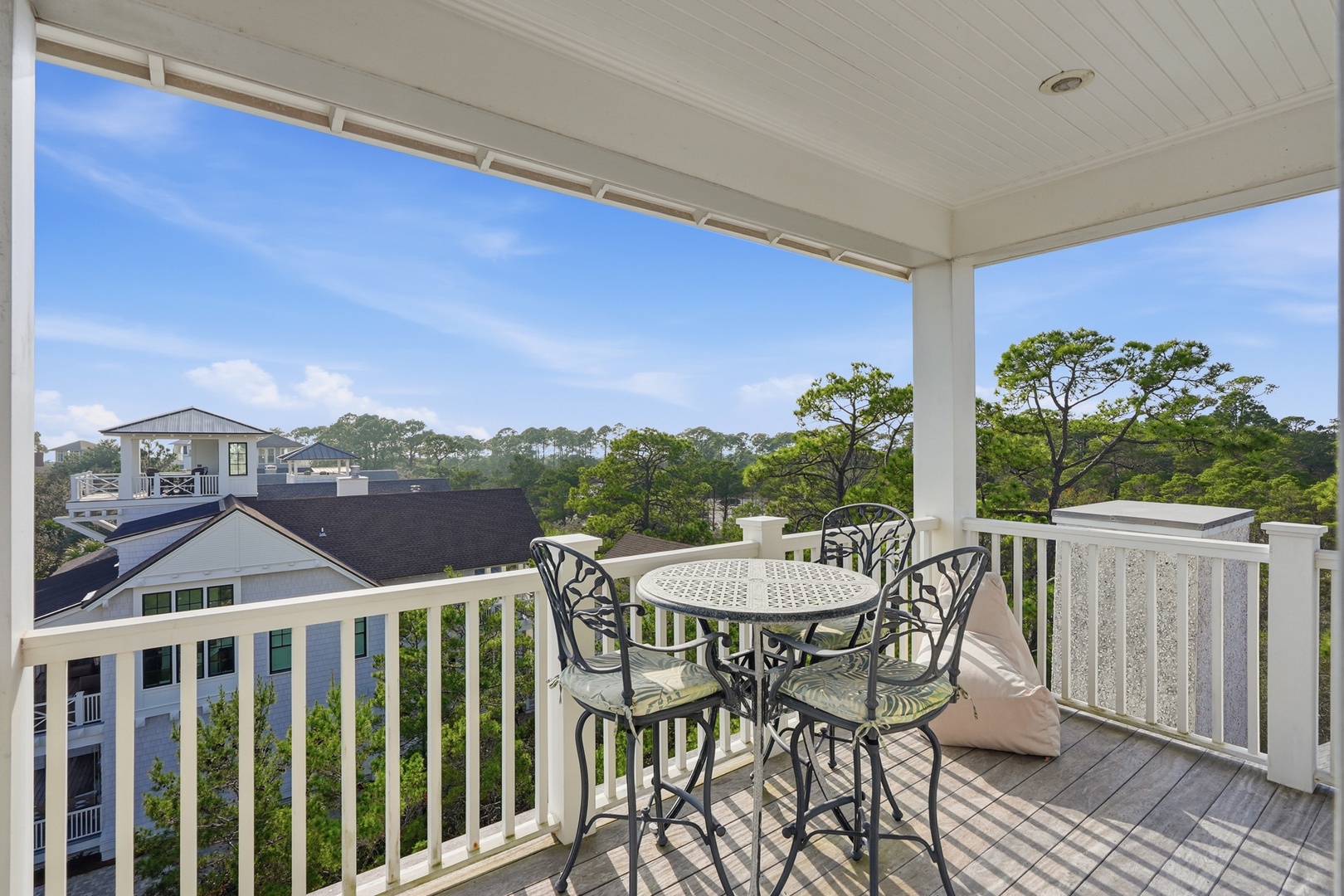 Elevated balcony overlooking neighborhood rooftops and tree canopy, furnished with bistro table and four chairs for outdoor dining.