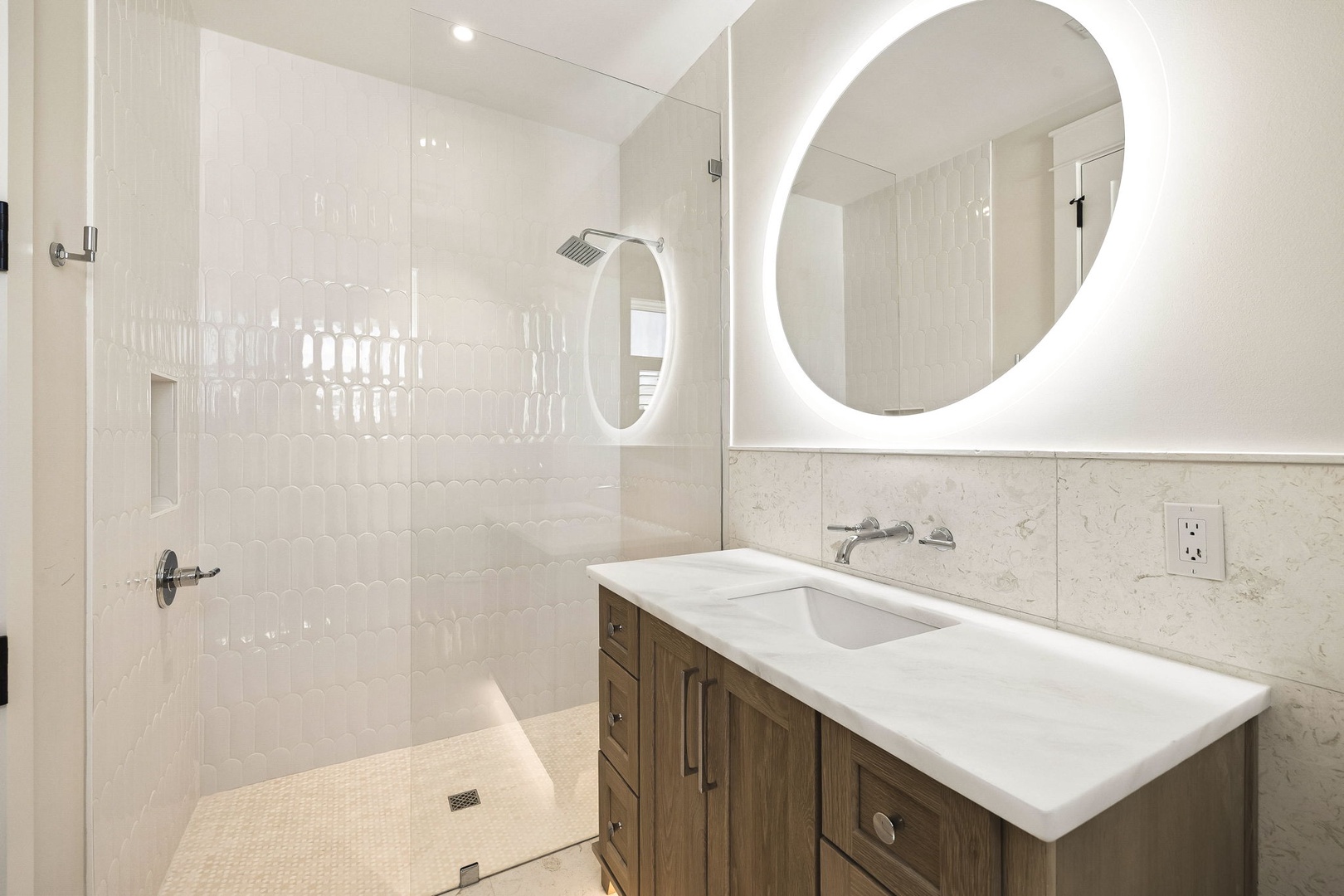 Step into a bright, modern bathroom where light marble surfaces and soft white tile create a spa-like sanctuary. Your walk-in shower awaits behind sleek glass, while the backlit mirror adds a serene glow to your morning rituals.