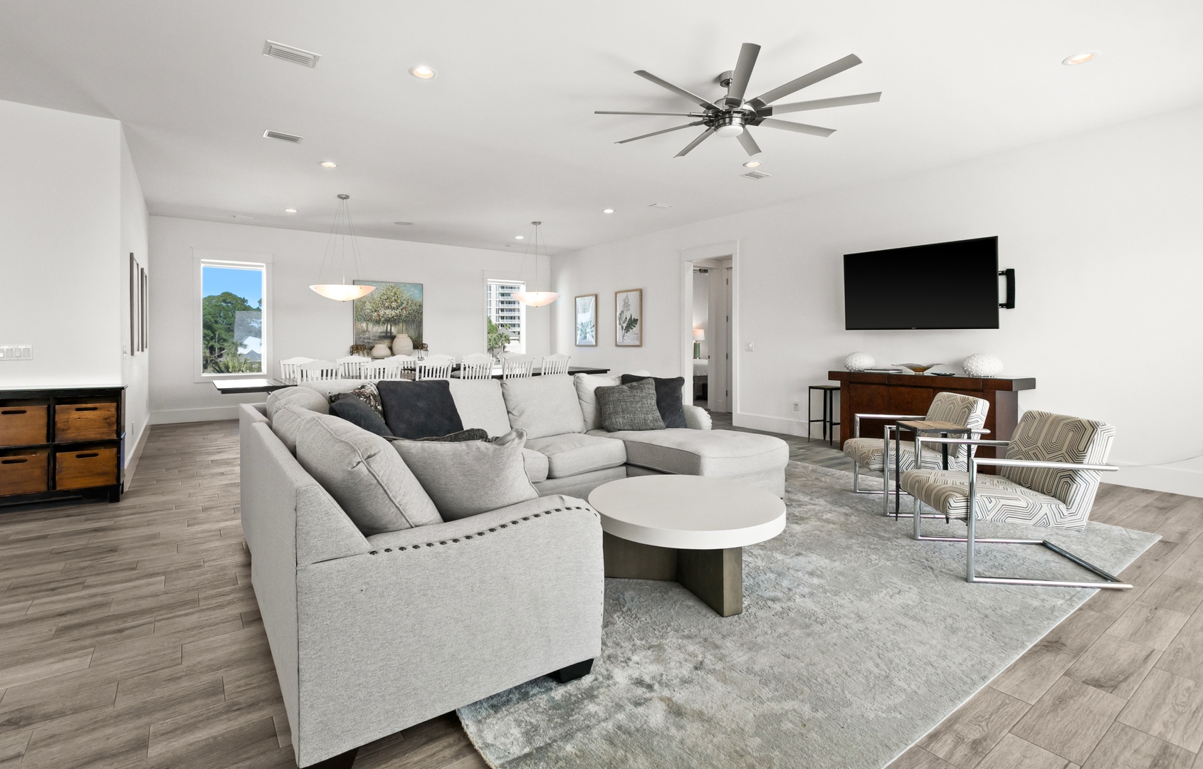 Spacious open-concept living area with plush seating, modern ceiling fans, and warm wood-look floors. Large windows flood the space with natural light, while a flat-screen TV and stylish furnishings create the perfect retreat.
