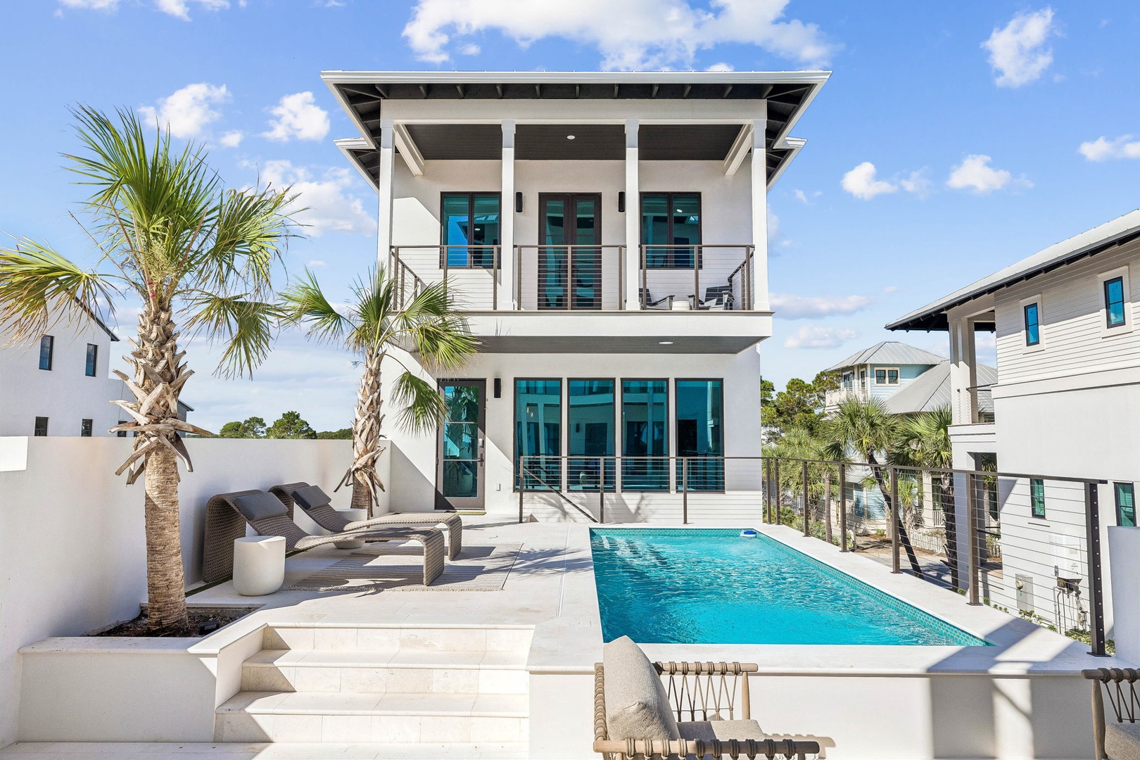 A modern coastal retreat featuring a pristine pool surrounded by palm trees and stylish lounge seating in a sun-drenched setting.
