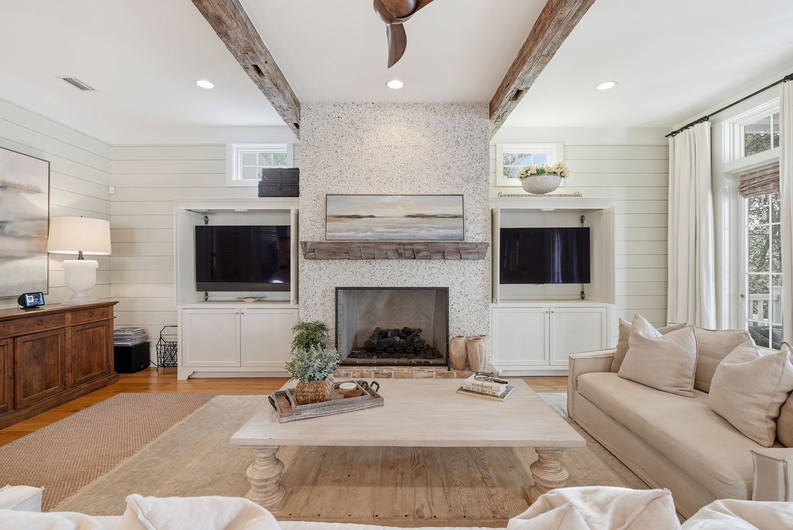Sink into the plush sofa and enjoy cozy evenings by the fireplace, flanked by built-in shelving and rustic wood beams overhead, all bathed in natural light from French doors.