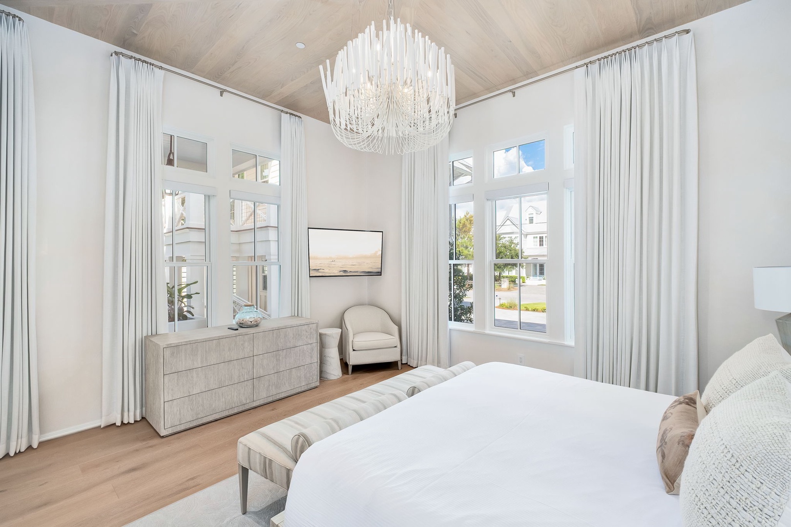 Serene bedroom retreat with vaulted ceilings, elegant chandelier, and tall windows flooding the space with natural light. Peaceful neighborhood views and coastal-inspired comfort await.