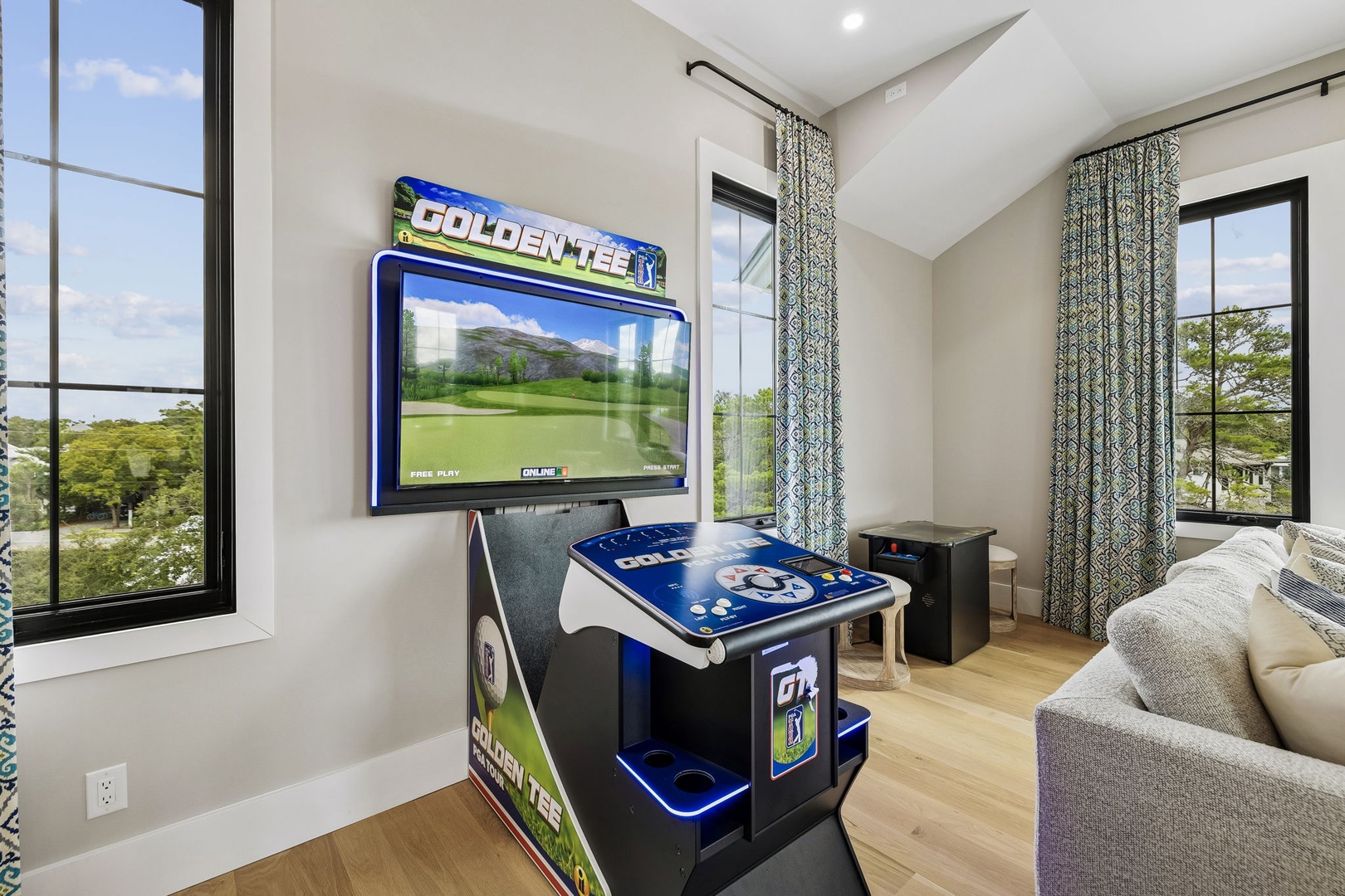 Your personal game room invites playful moments with a classic arcade setup and soaring vaulted ceilings. Natural light pours in through large windows framing lush greenery, while plush seating ensures cozy relaxation between turns.