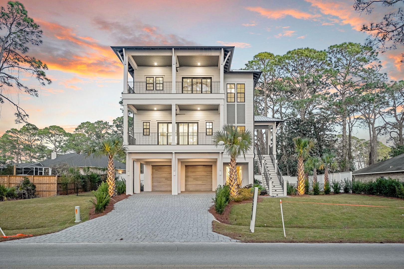 Modern three-story vacation home featuring coastal architecture with palm-lined driveway and spacious balconies set among mature trees.